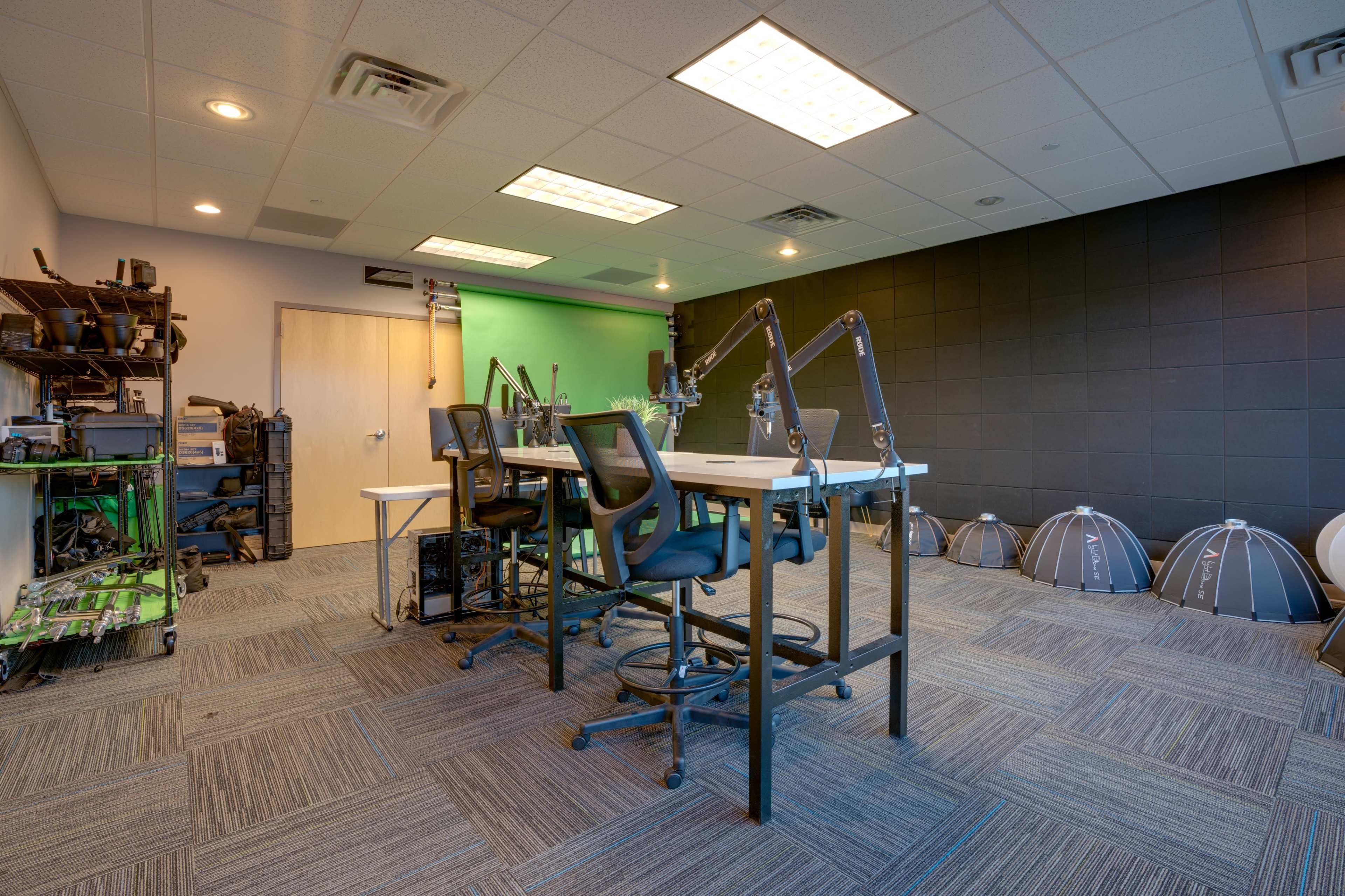 A modern studio space with a green screen, two desks with adjustable stools, and various equipment organized along the walls.
