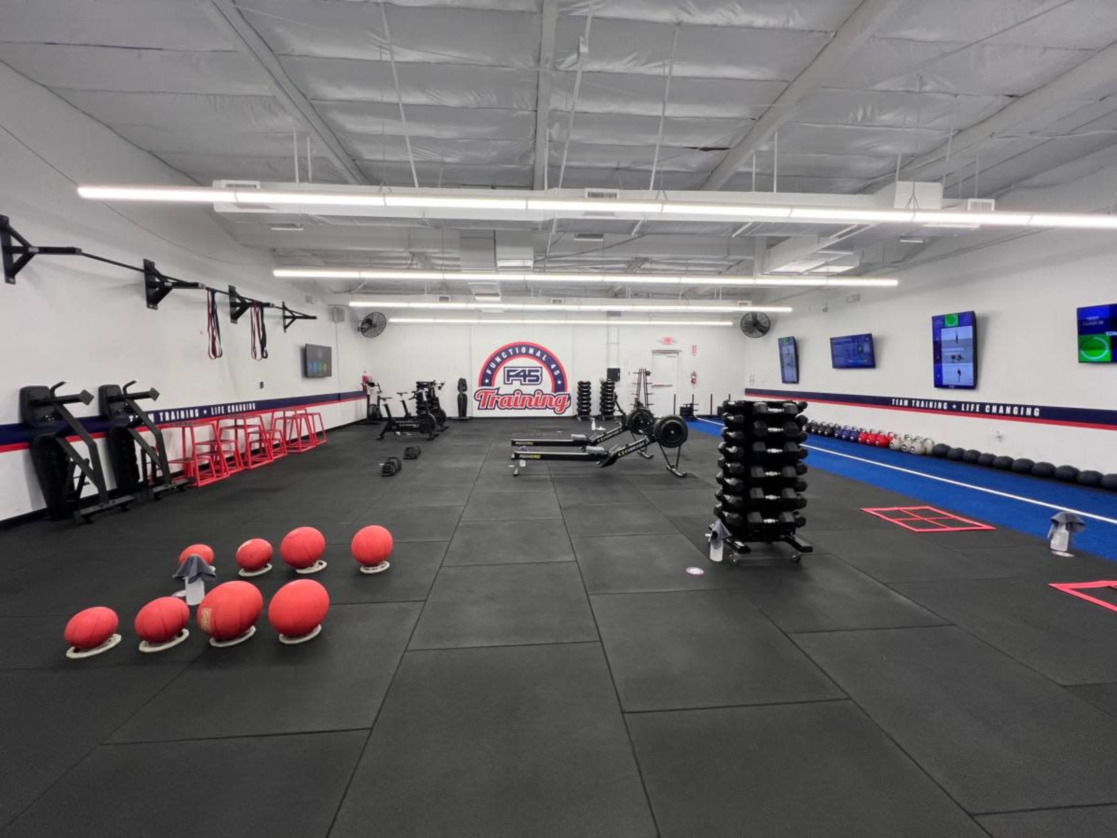 The image shows a gym interior with various fitness equipment, including weights, benches, and exercise balls arranged across a black rubber floor.