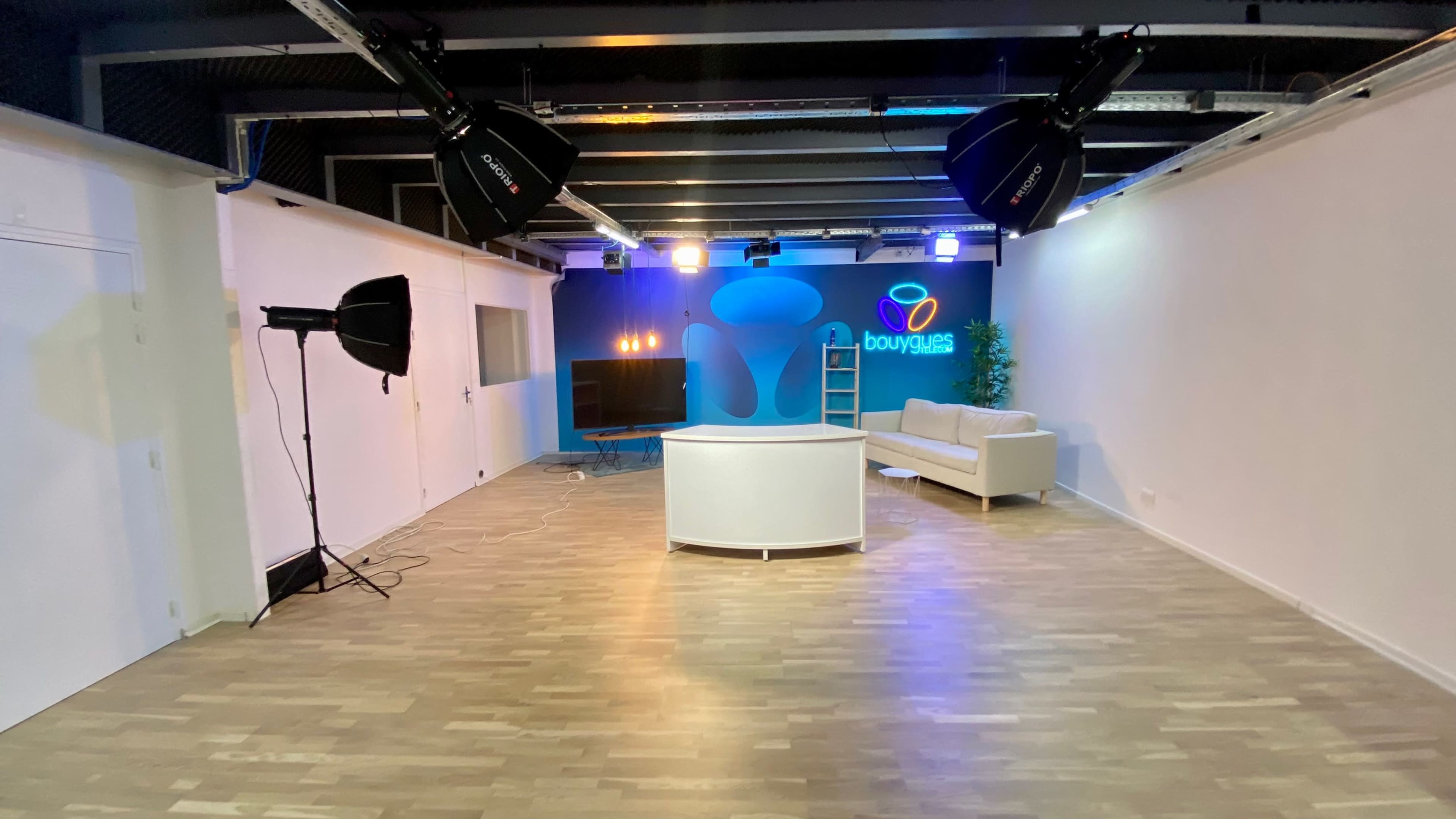 A modern studio space with a white central reception desk, a couch, and professional lighting equipment set up against a backdrop featuring a colorful logo.