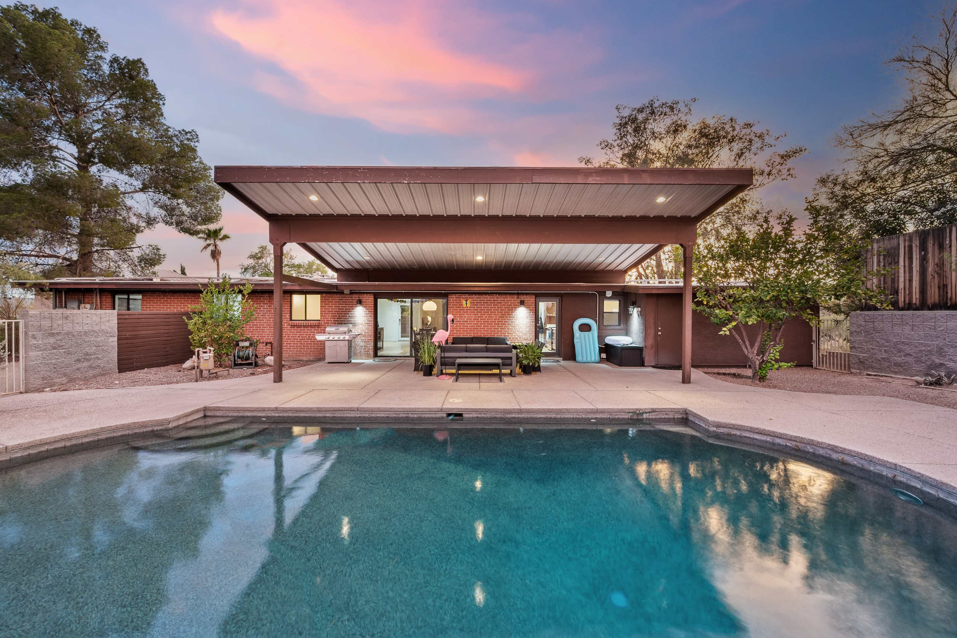 A patio area with a swimming pool is shown behind a house, featuring a covered seating space and landscaped plants under a colorful sunset sky.