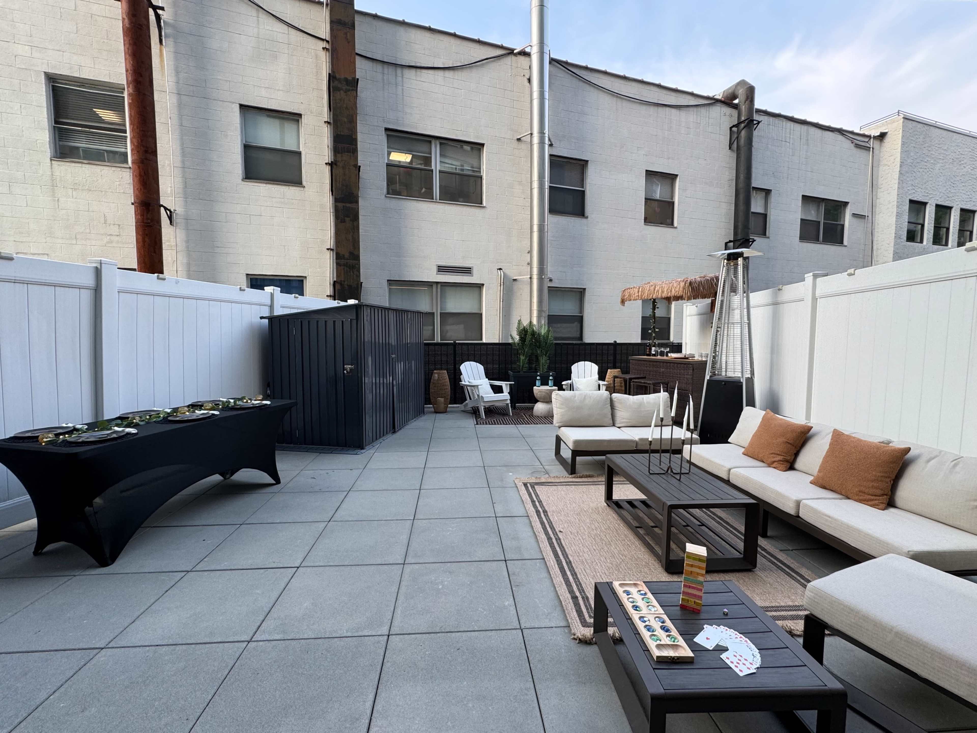 The image shows a patio area with a seating arrangement, a table set for snacks, and various lounge chairs against a backdrop of brick buildings.