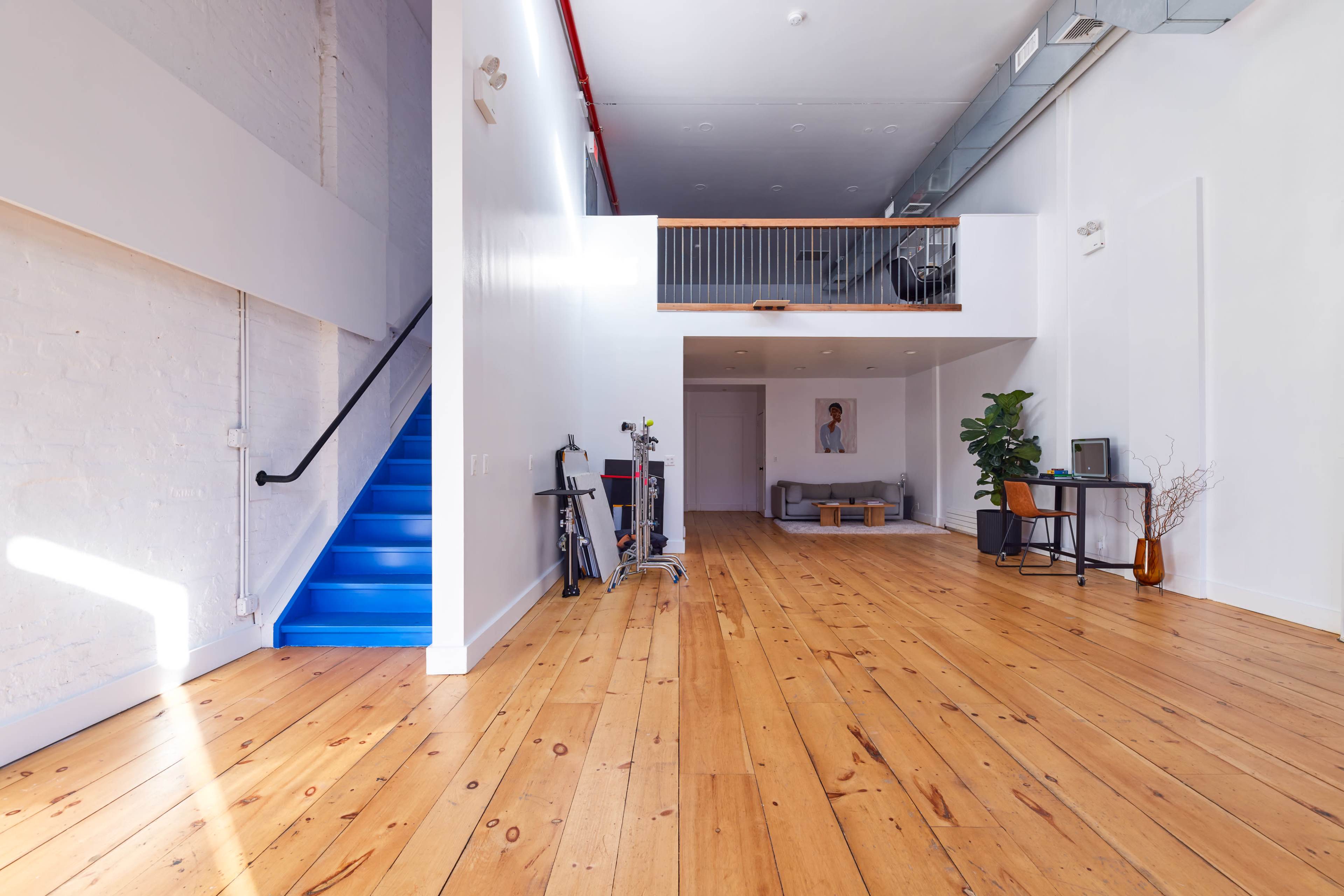 The image shows a bright, open interior space with wooden floors, a staircase painted blue, and minimalist furniture.