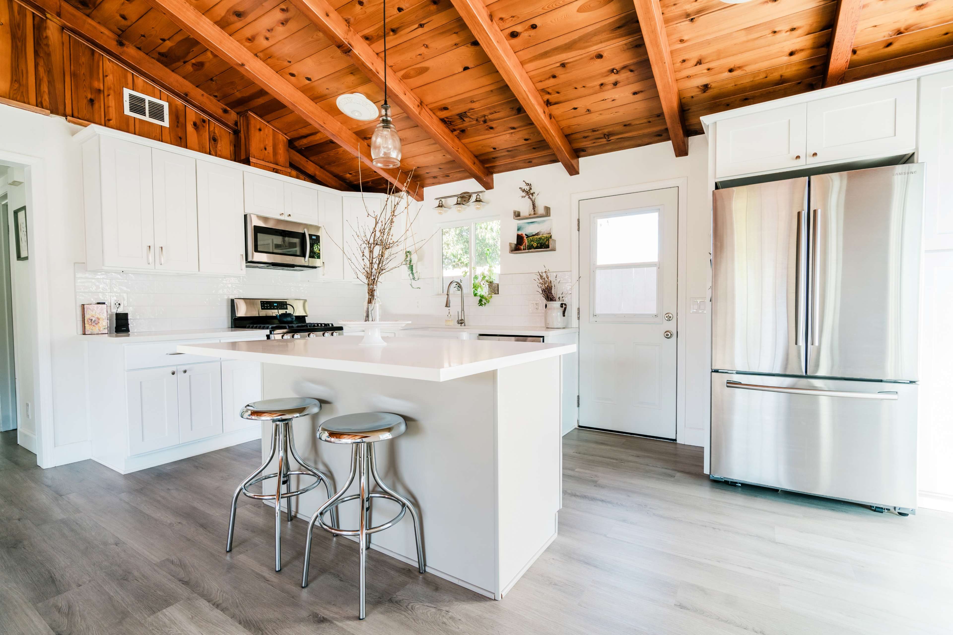 The image shows a modern kitchen with white cabinetry, a central island with two barstools, and wooden beams on the ceiling.