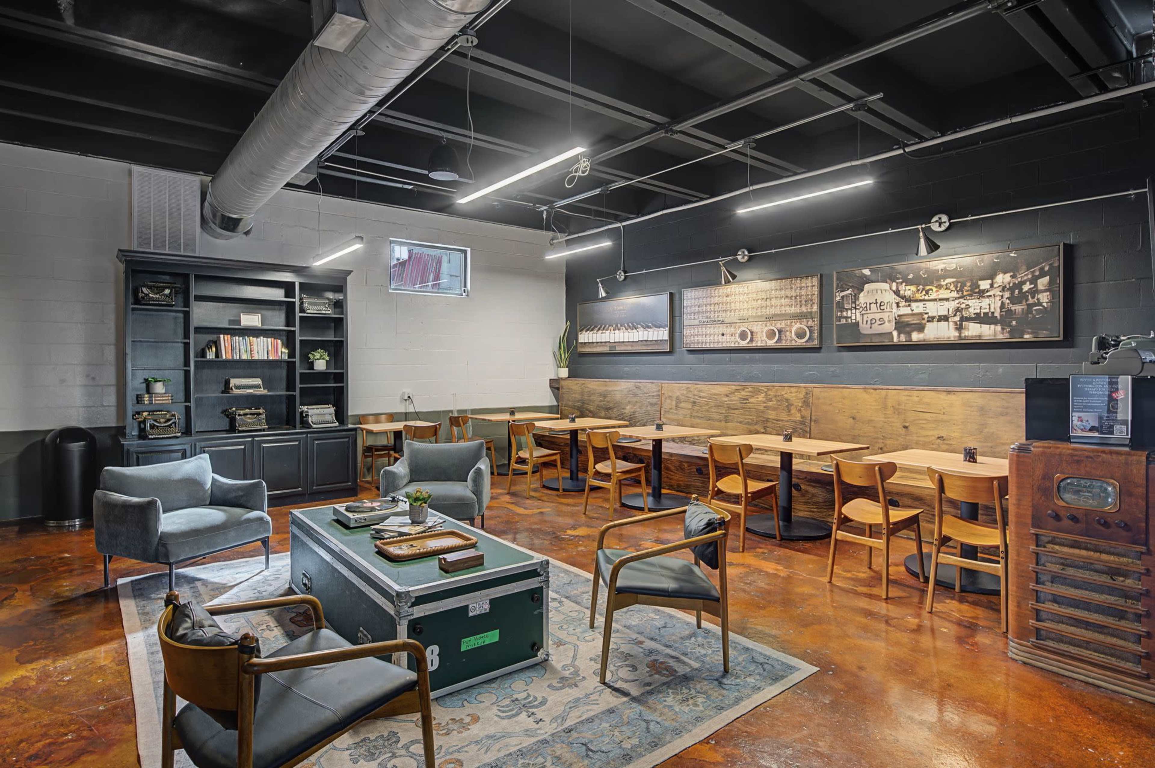 The image shows a modern workspace featuring a mix of seating arrangements, including chairs and tables, against a backdrop of dark walls and wooden accents.