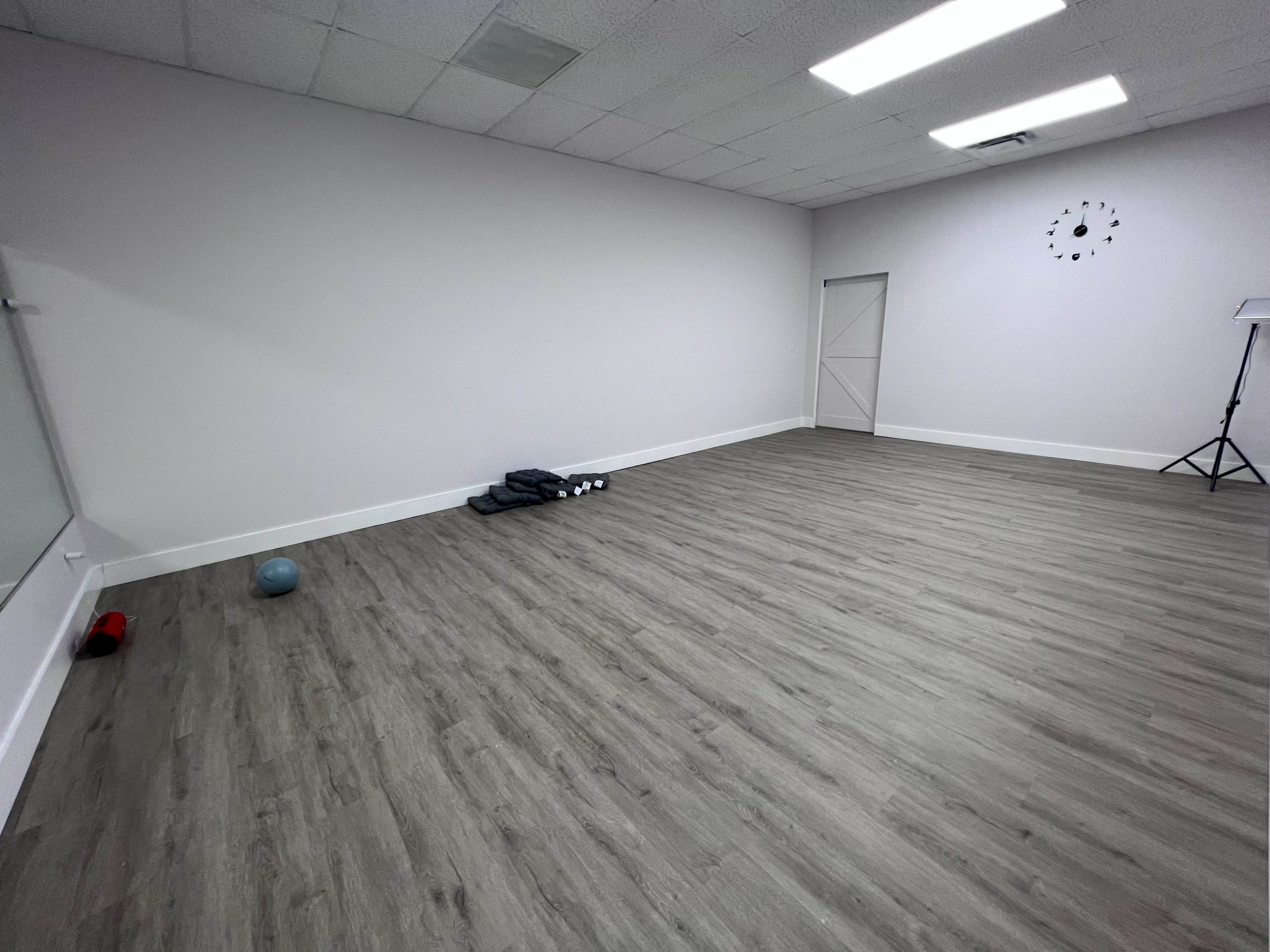 An empty exercise studio with a light-colored floor, a large mirror on one wall, and fitness equipment neatly arranged in one corner.