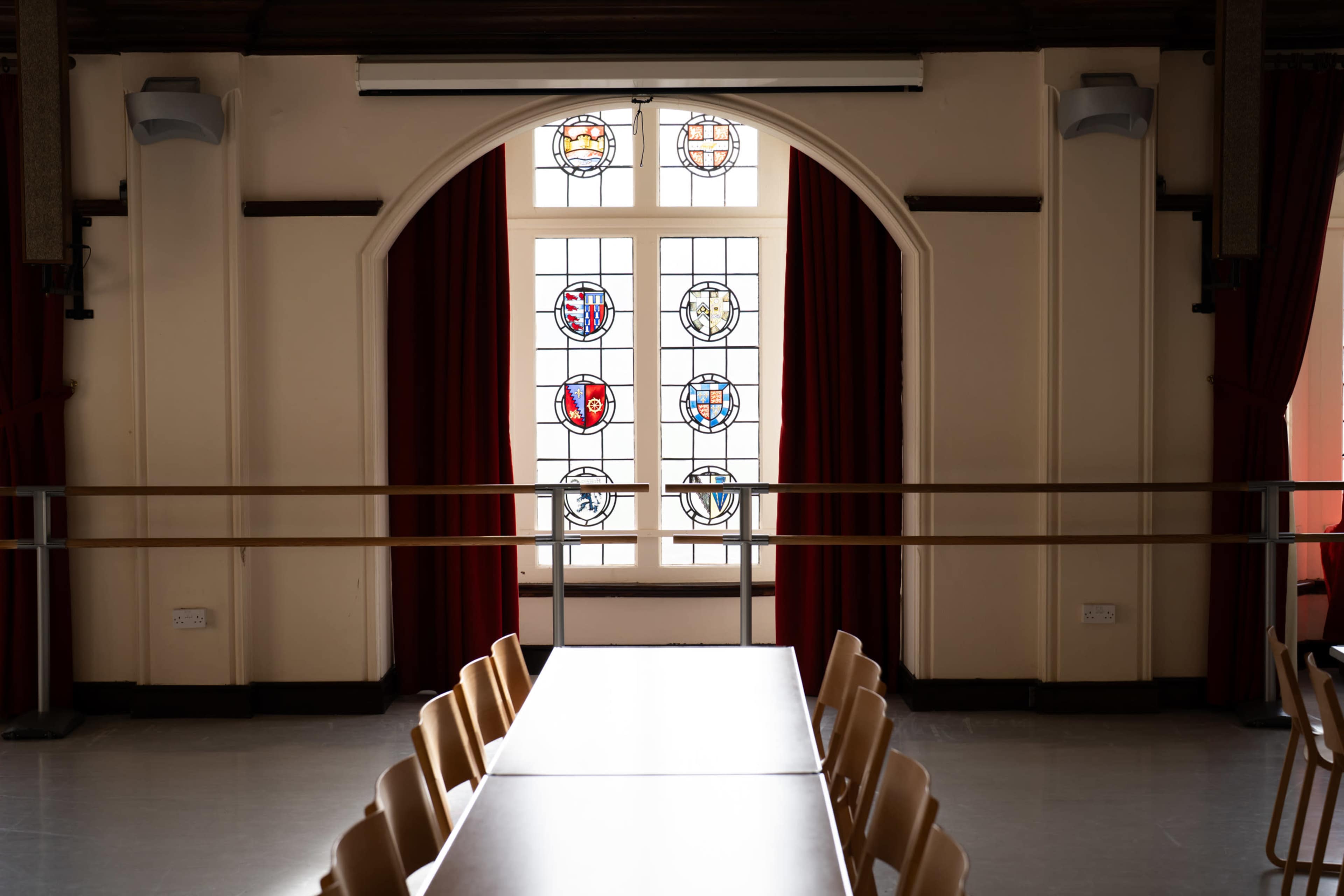 The image shows a spacious indoor room with a long table and stained glass windows featuring various designs in the background.