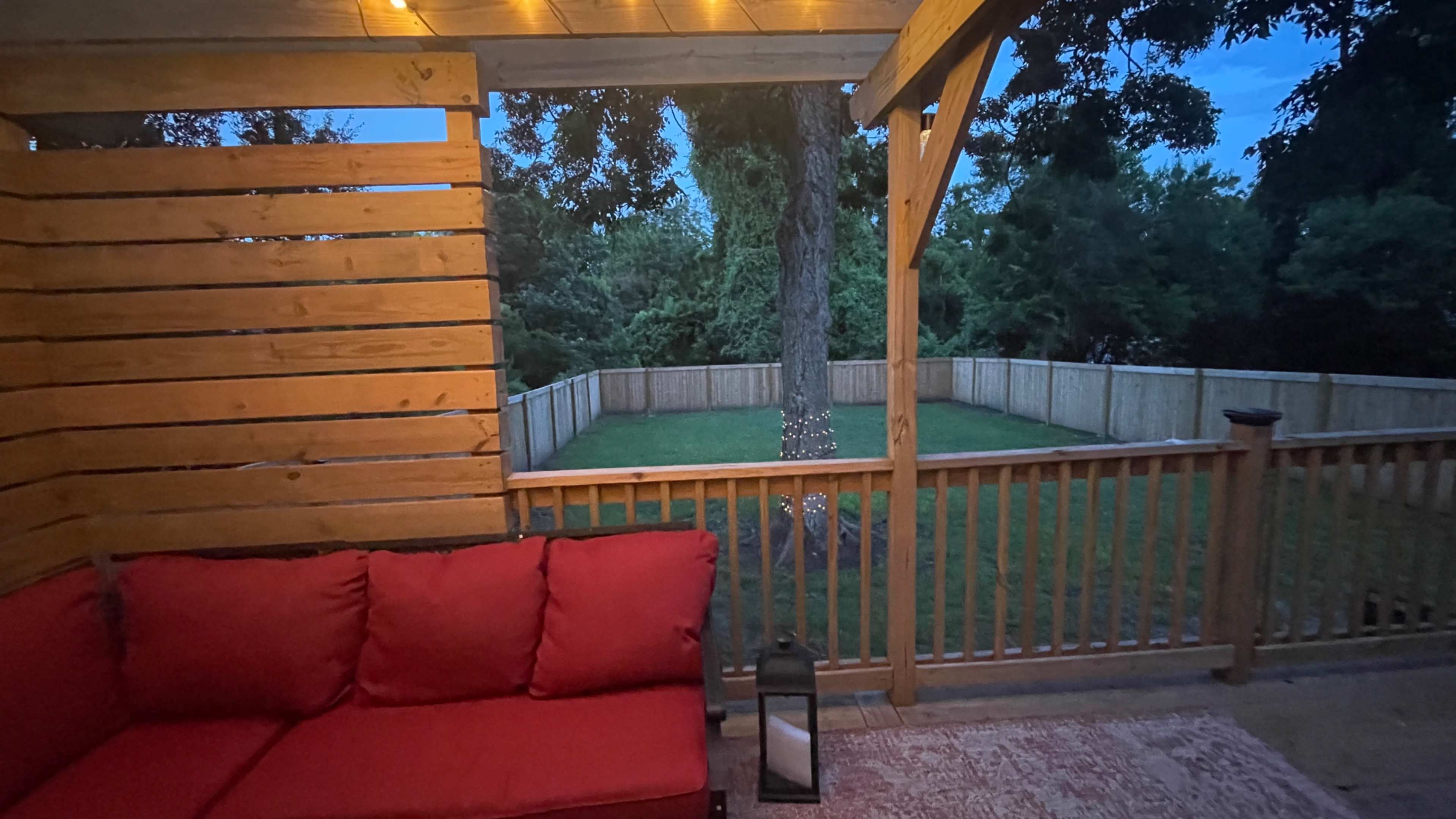 A cozy outdoor seating area features a red couch, lantern, and a view of a fenced yard with a tree.