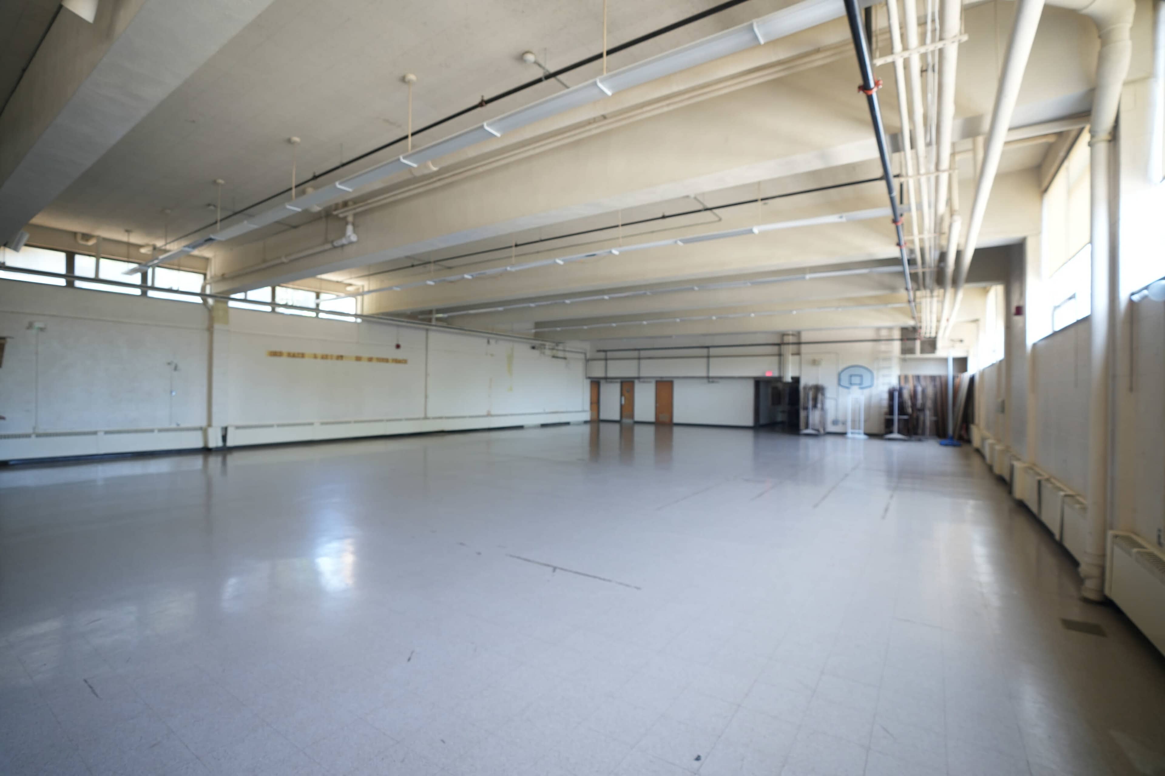 The image shows a large, empty gymnasium with high ceilings and multiple windows.