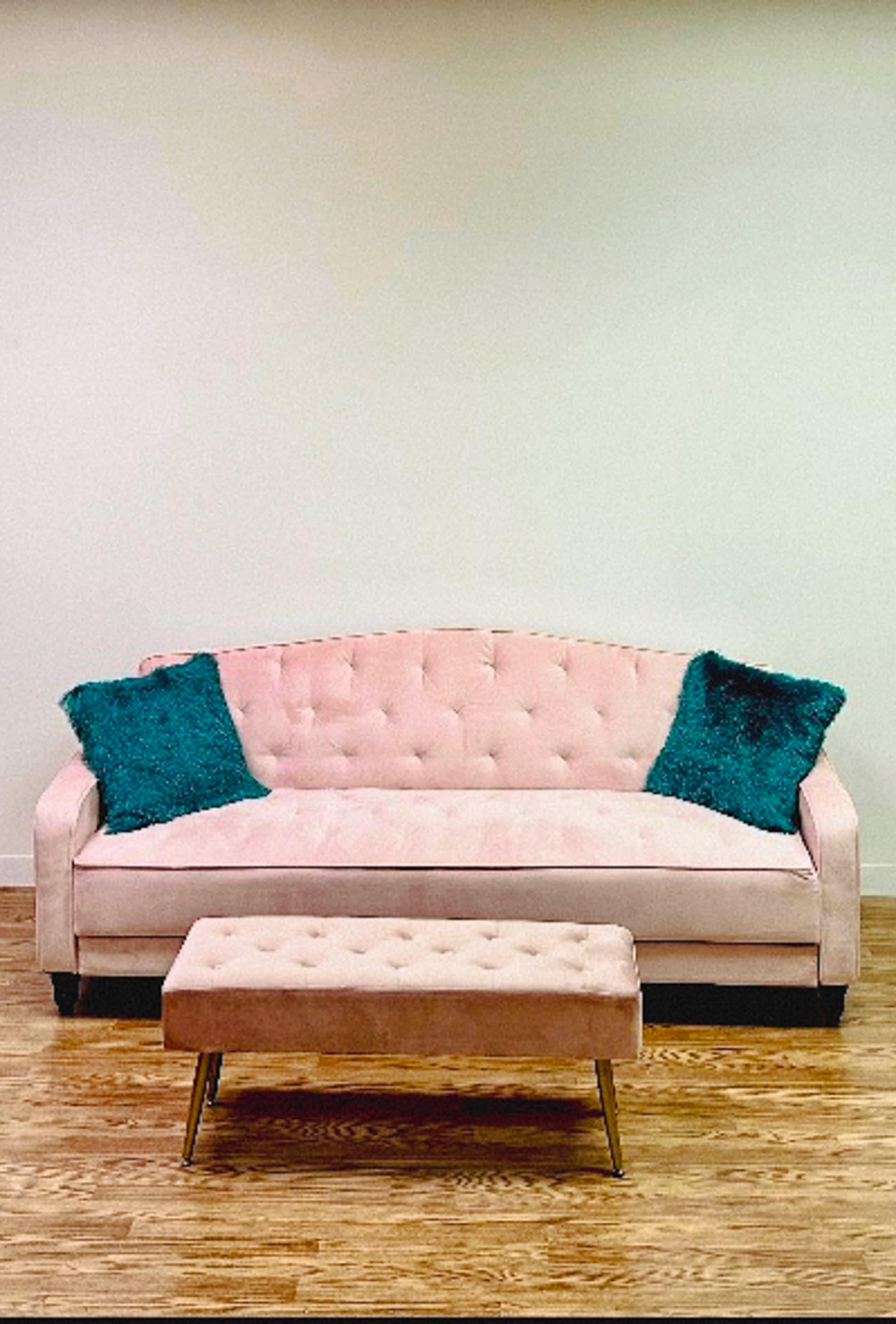 A pink tufted sofa with two teal pillows and a matching ottoman sits on a wooden floor against a plain wall.