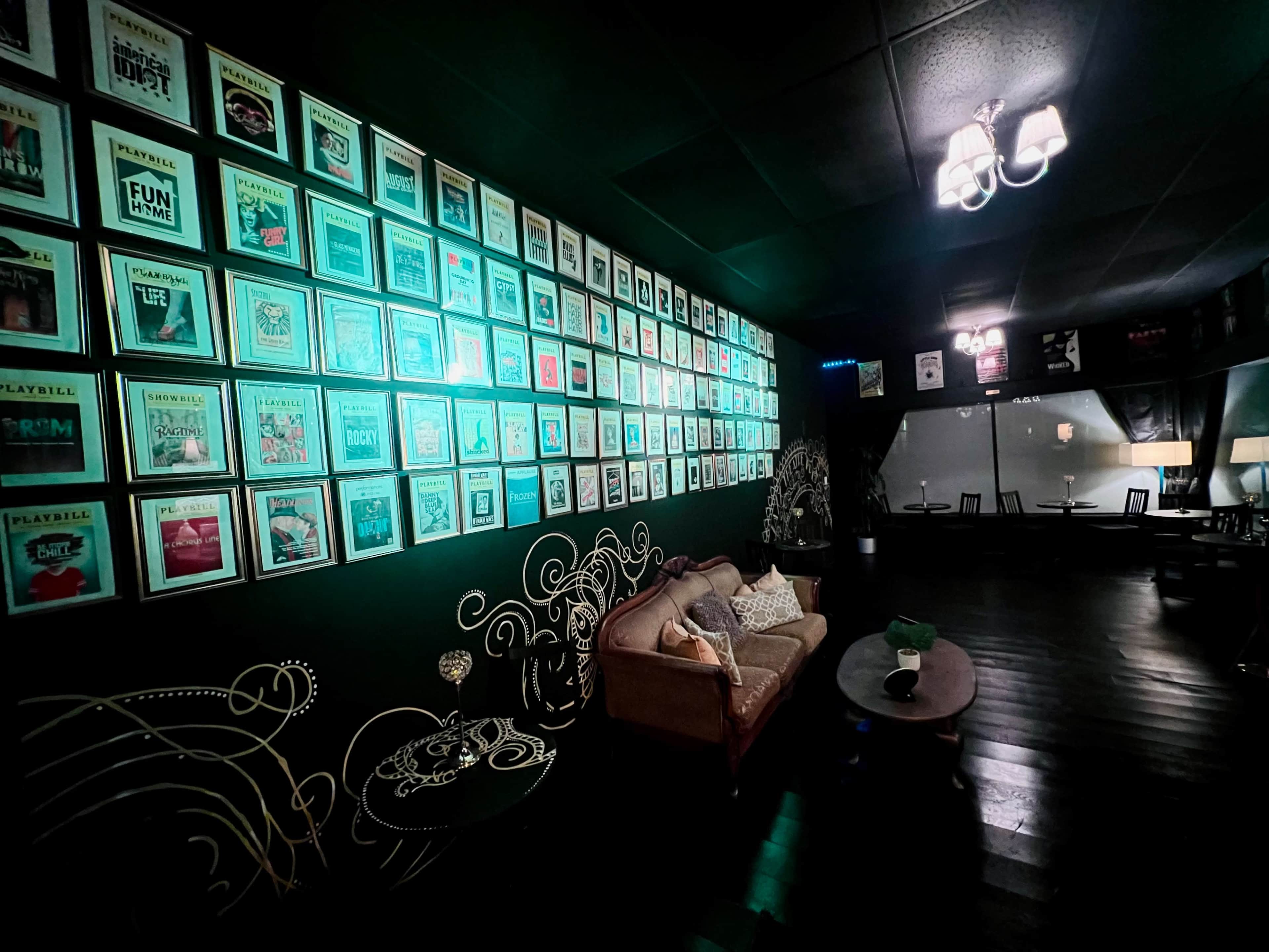 The image shows a dimly lit lounge with a wall decorated with framed playbills and a seating area featuring a sofa and coffee table.