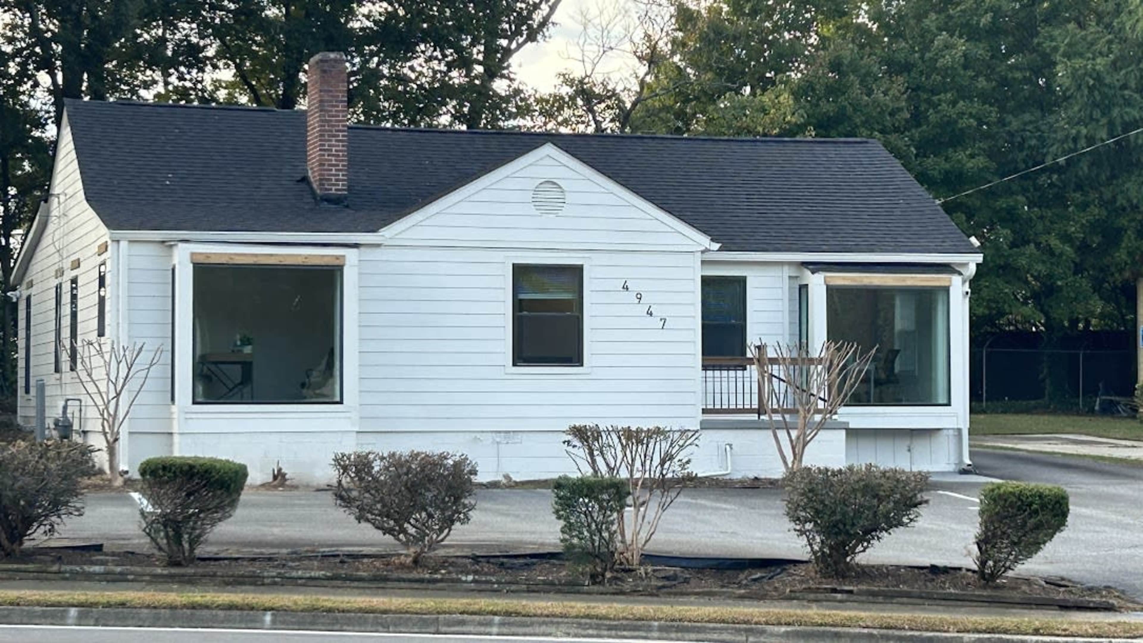 A single-story white house with a black roof and front porch, situated on a gravel driveway.