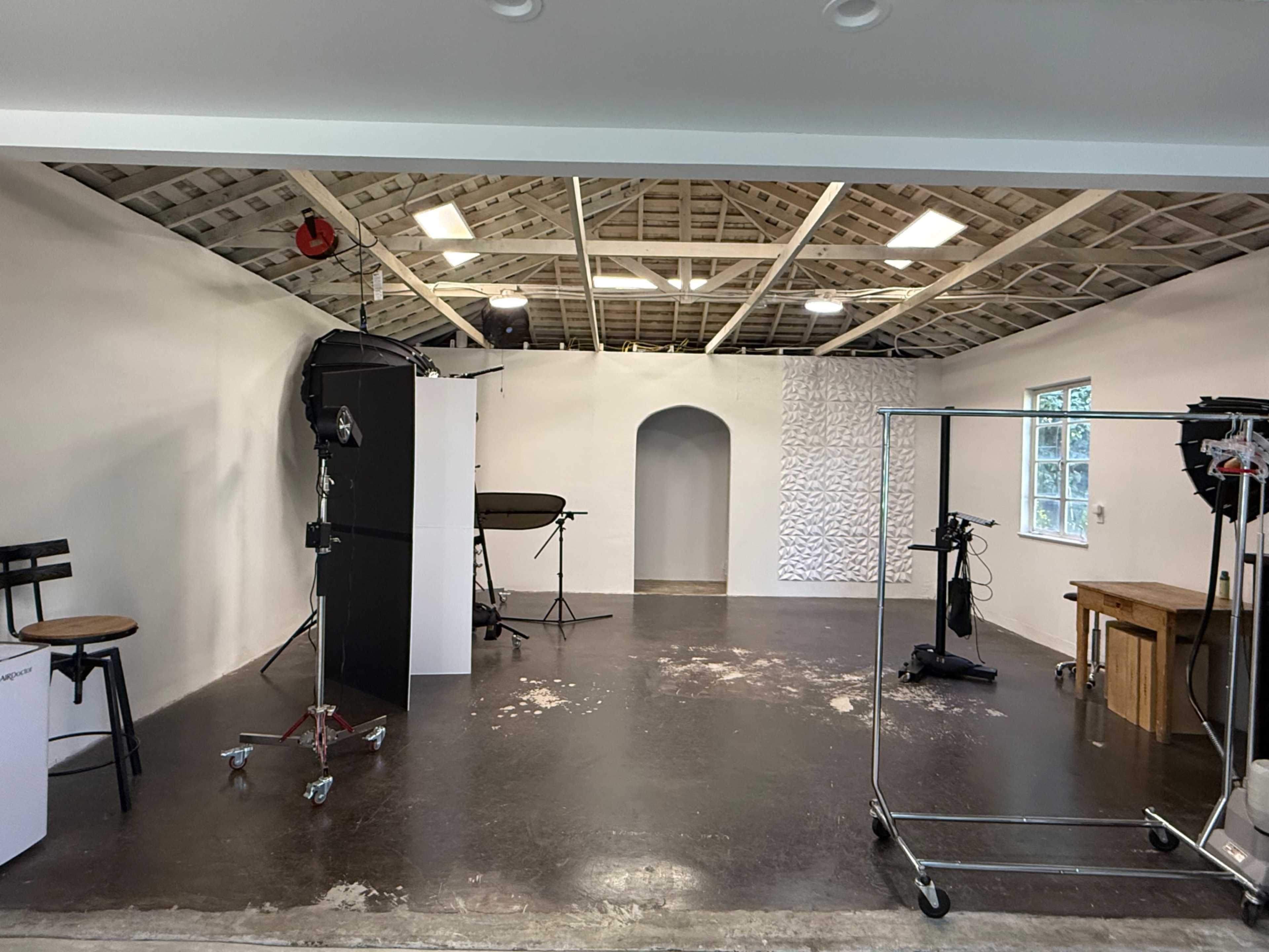 The image shows a spacious photo studio with a wooden floor, white walls, various lighting equipment, and a couple of props including a chair and a table.