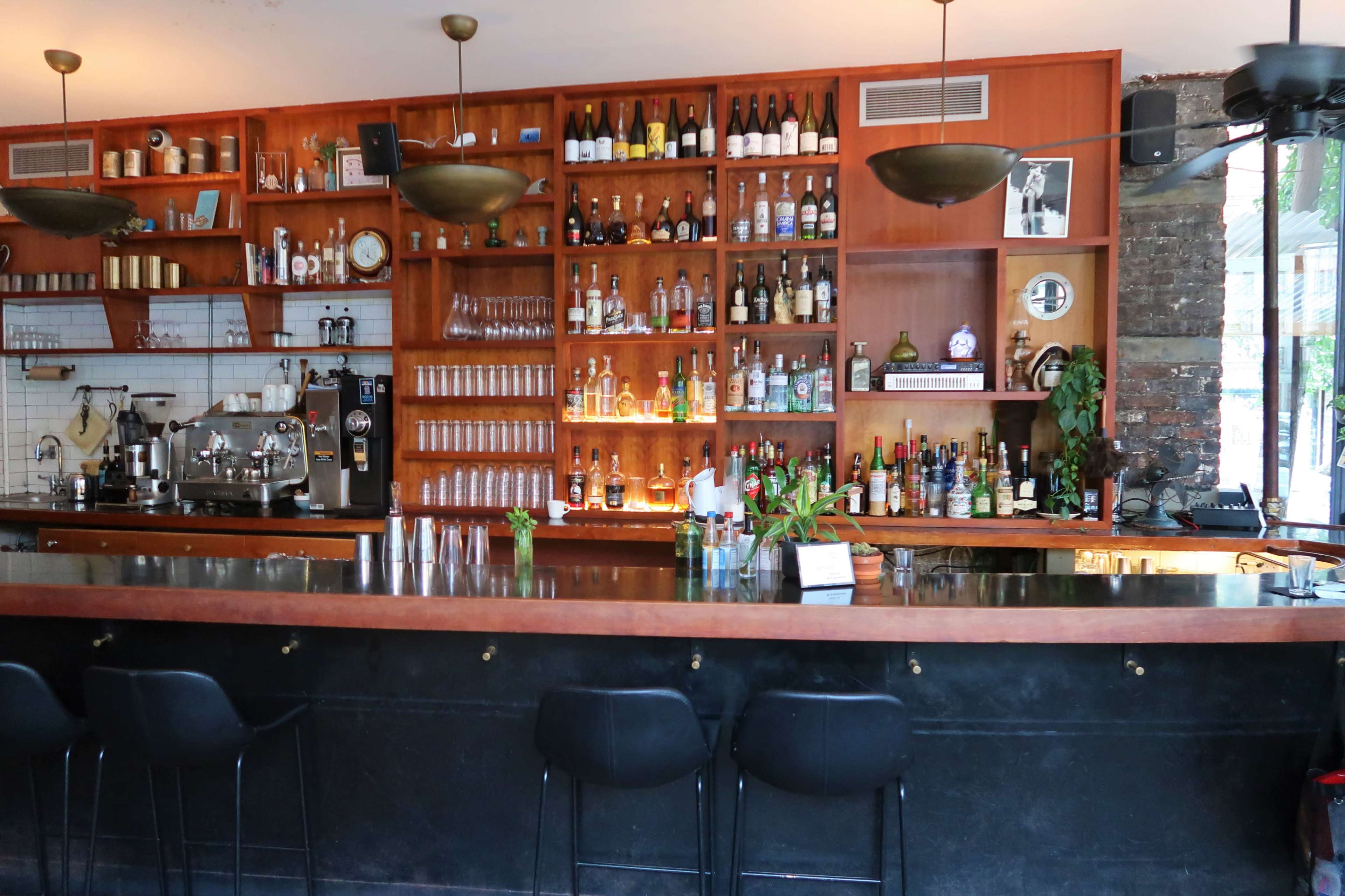 A well-organized bar with a wooden shelving unit displaying various bottles and glassware, alongside a sleek black counter and a few stools.
