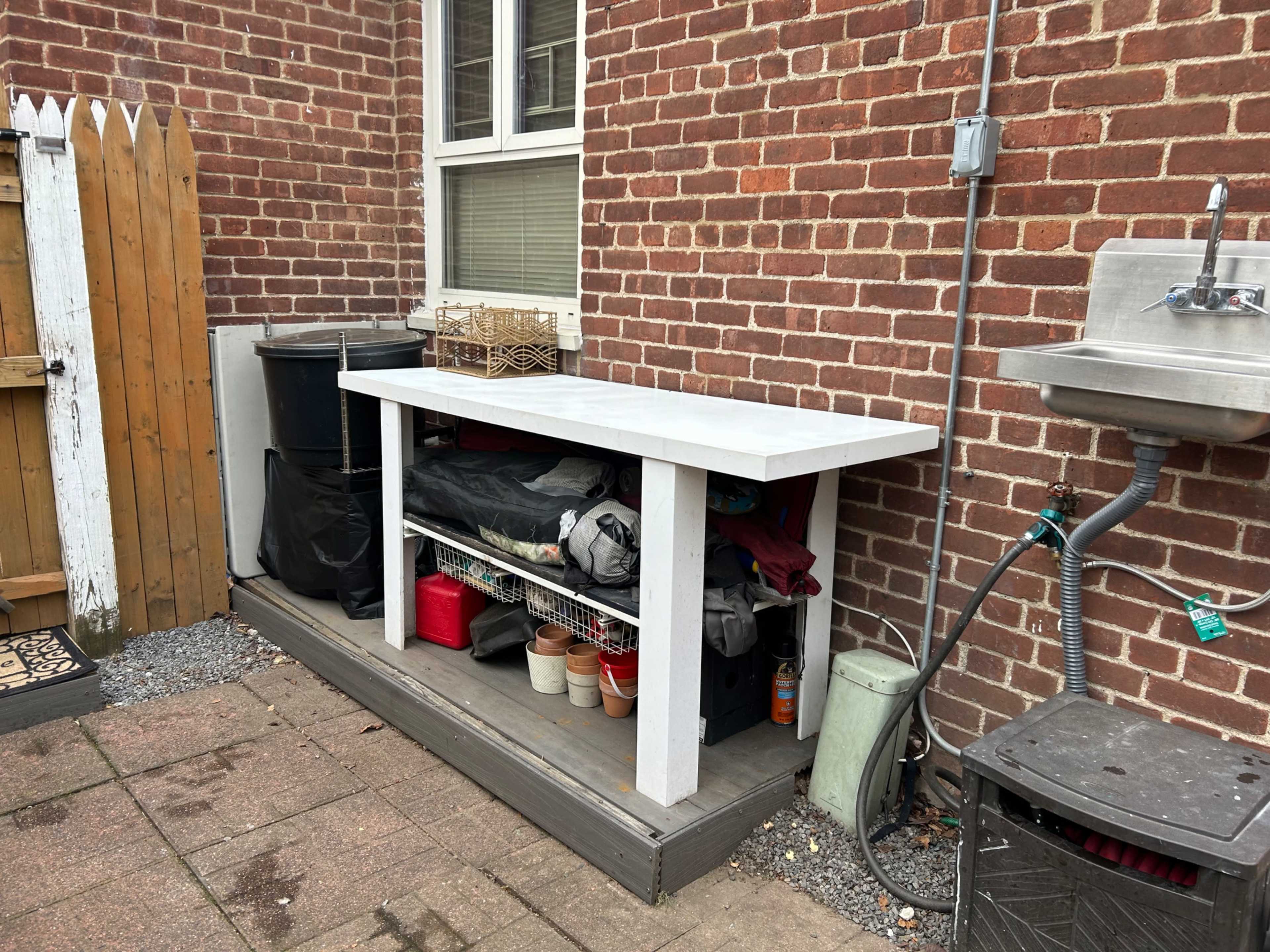A white table with storage underneath is situated against a brick wall, accompanied by a sink and various containers.