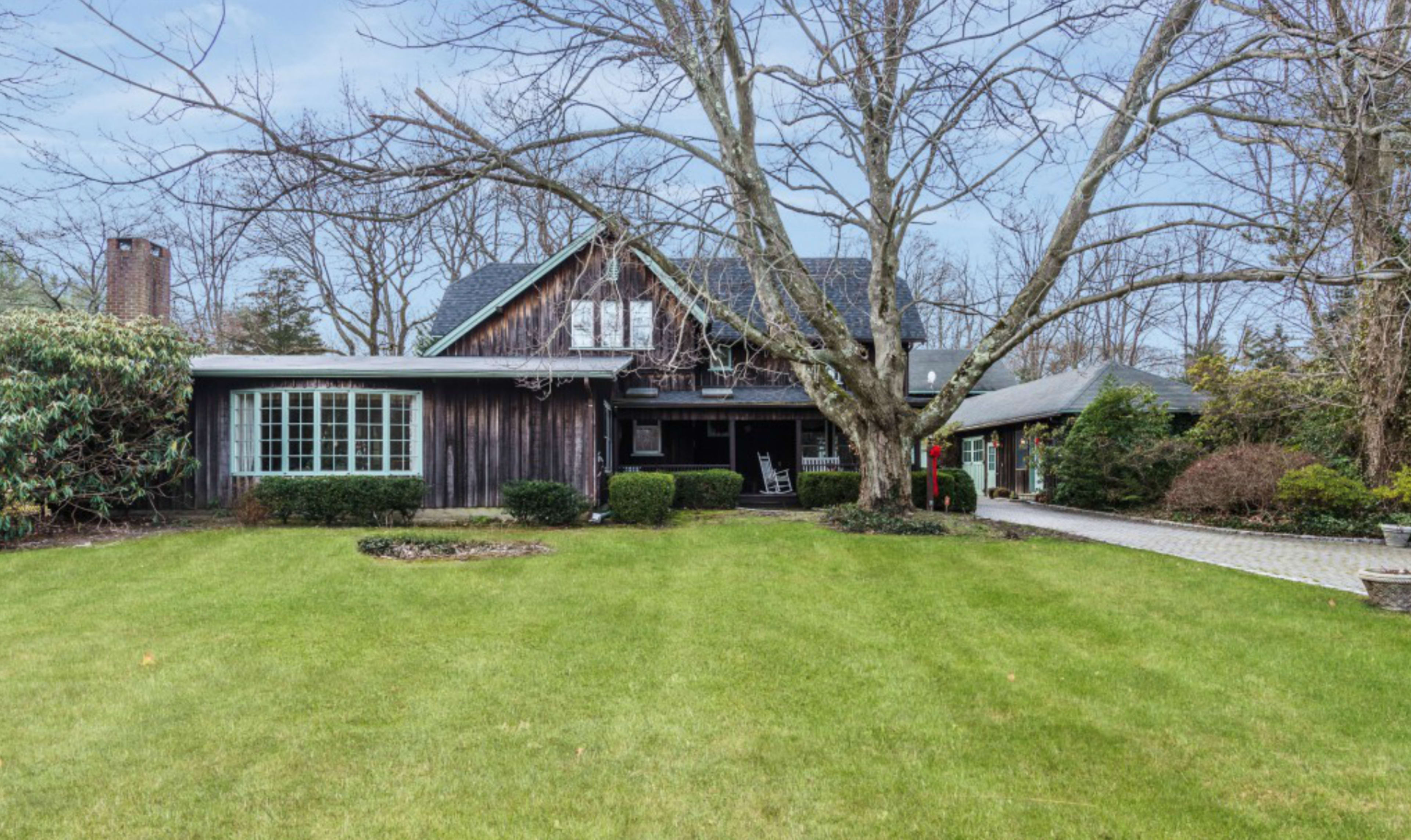 A two-story wooden house with a large front lawn and bare trees is surrounded by landscaped shrubs.