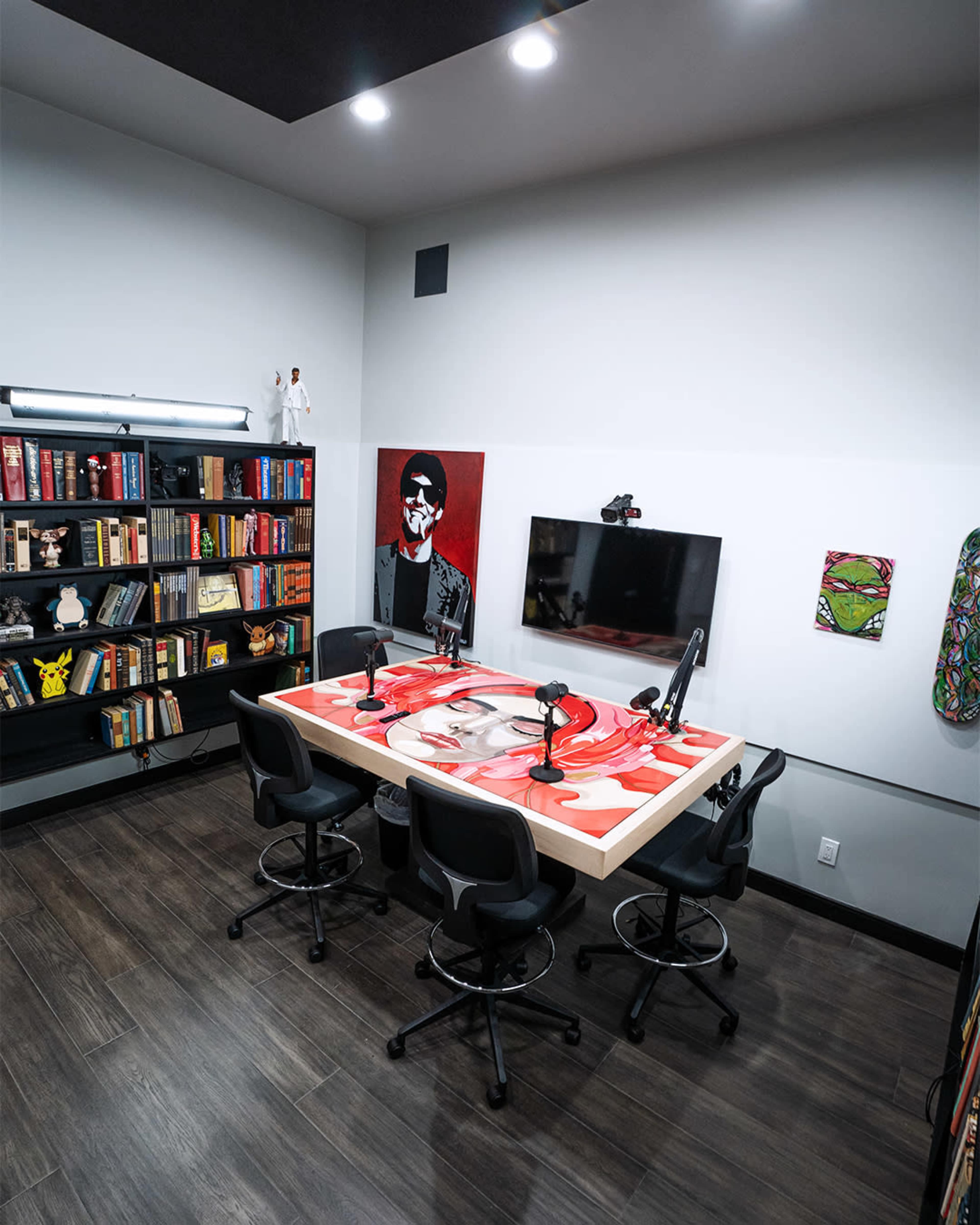 A modern podcast studio with a large table, four microphone setups, a wall-mounted television, and bookshelves filled with various books and decor.