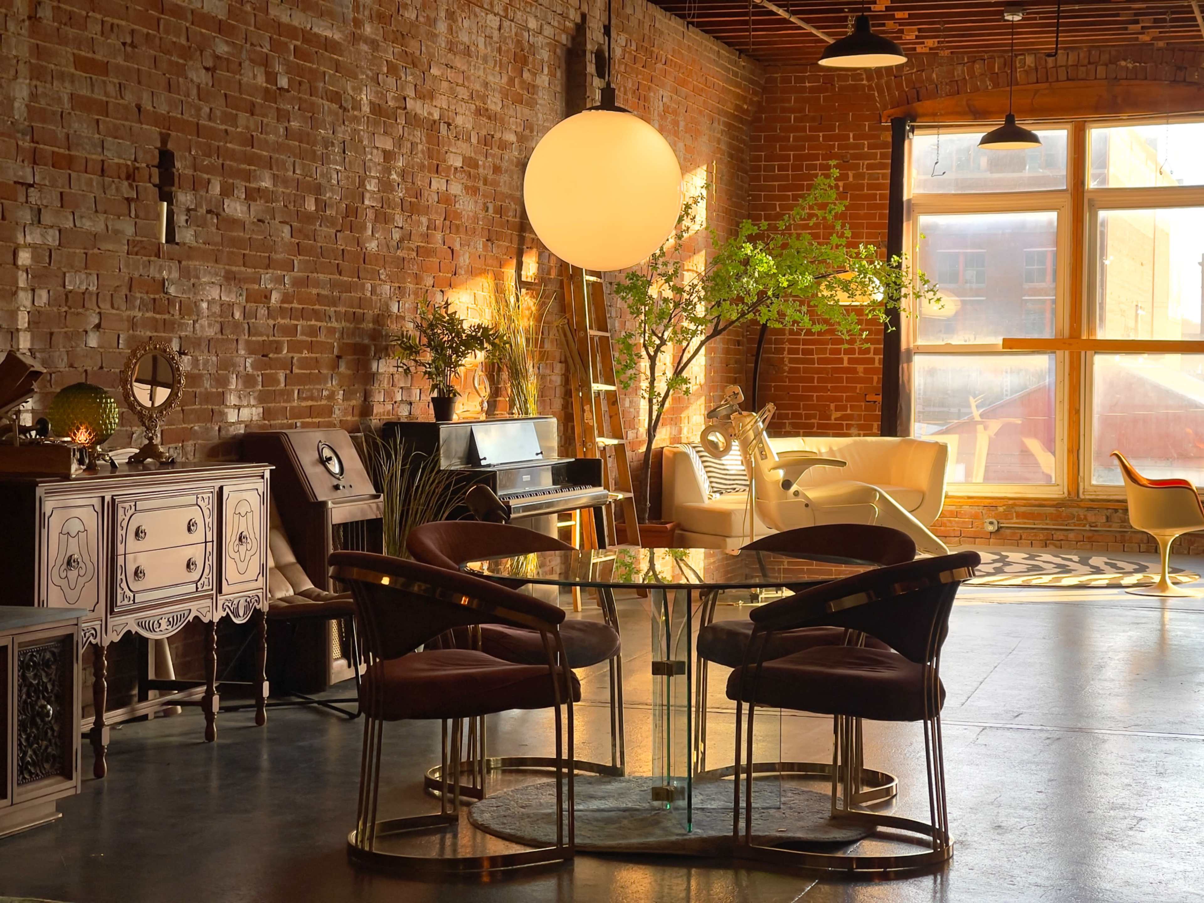 The image shows a spacious, industrial-style loft with exposed brick walls, large windows, and a mix of modern and vintage furniture.