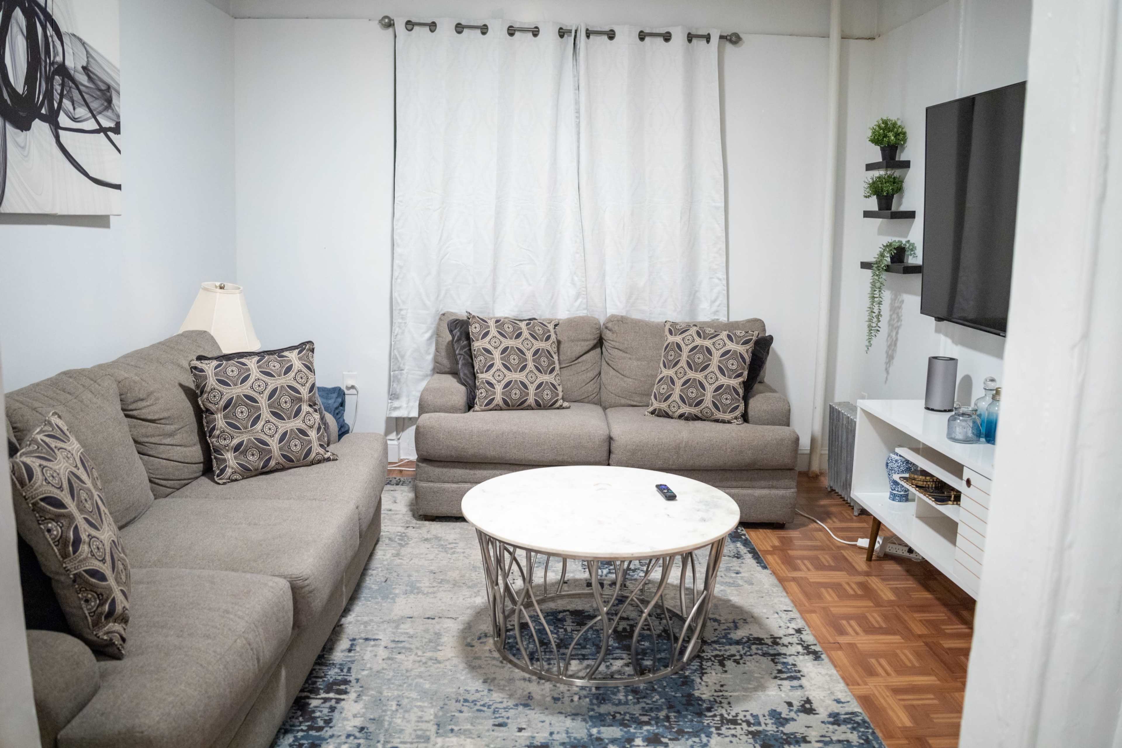 A living room with two gray sofas arranged around a round coffee table, set on a patterned rug, with a television mounted on the wall and curtains covering a window.