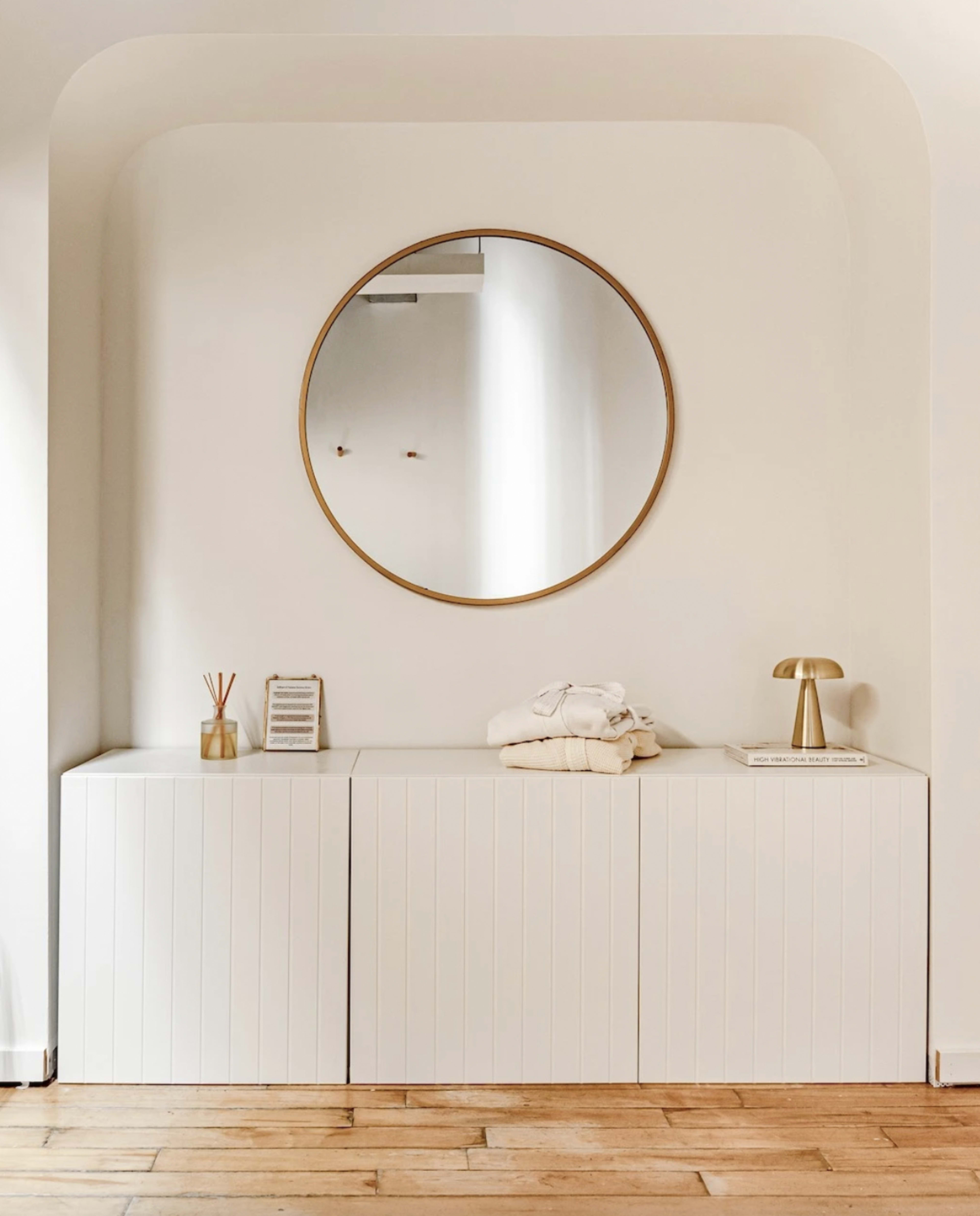 A round mirror is centered above a white storage cabinet, with a lamp, a towel, and decorative items arranged on top.