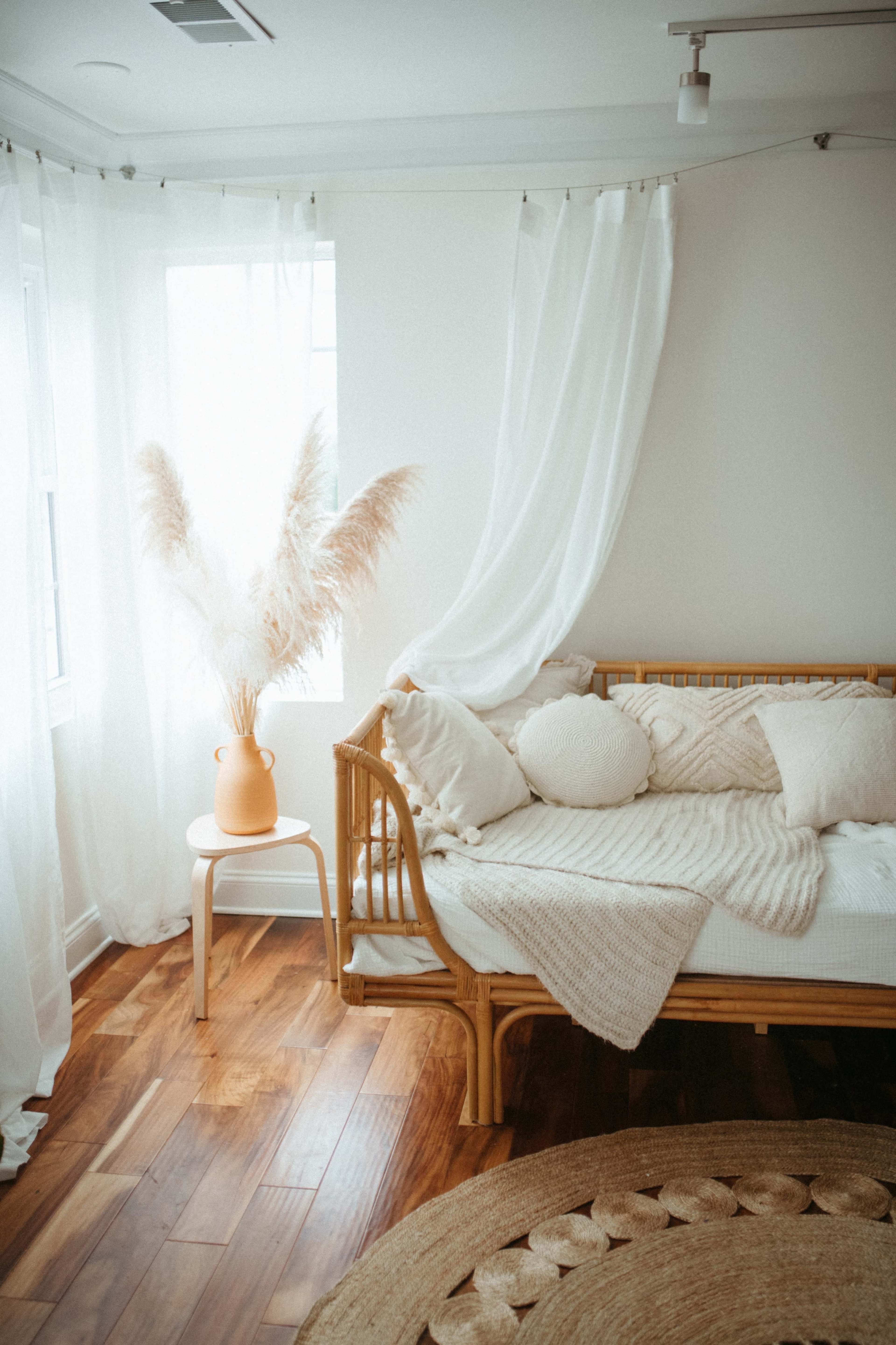 A bright, minimalist bedroom featuring a wooden daybed with neutral pillows, a large plant in a terracotta pot, and light-colored curtains.