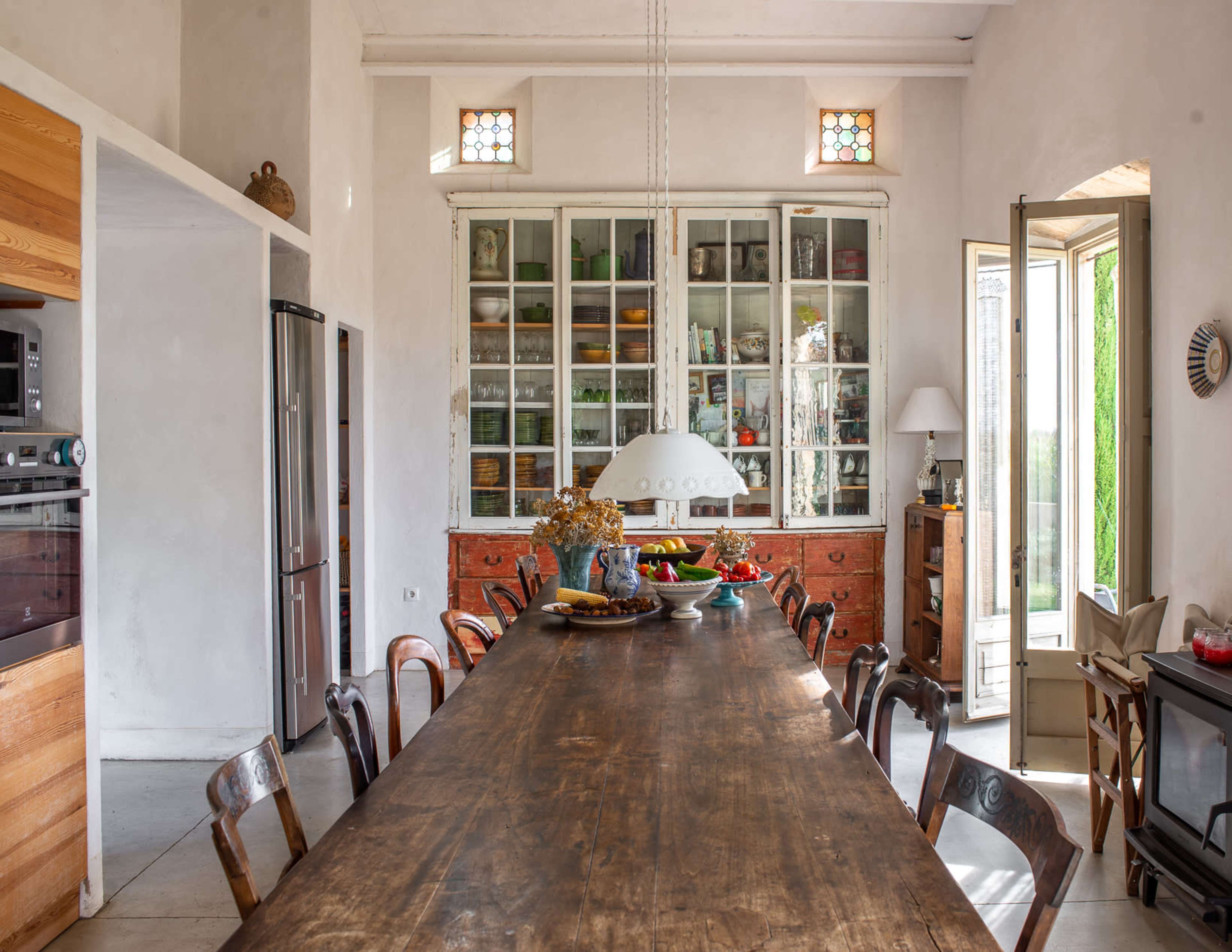 A spacious kitchen with a long wooden table, large glass cabinets displaying various dishes, and an open door leading to an outdoor area.