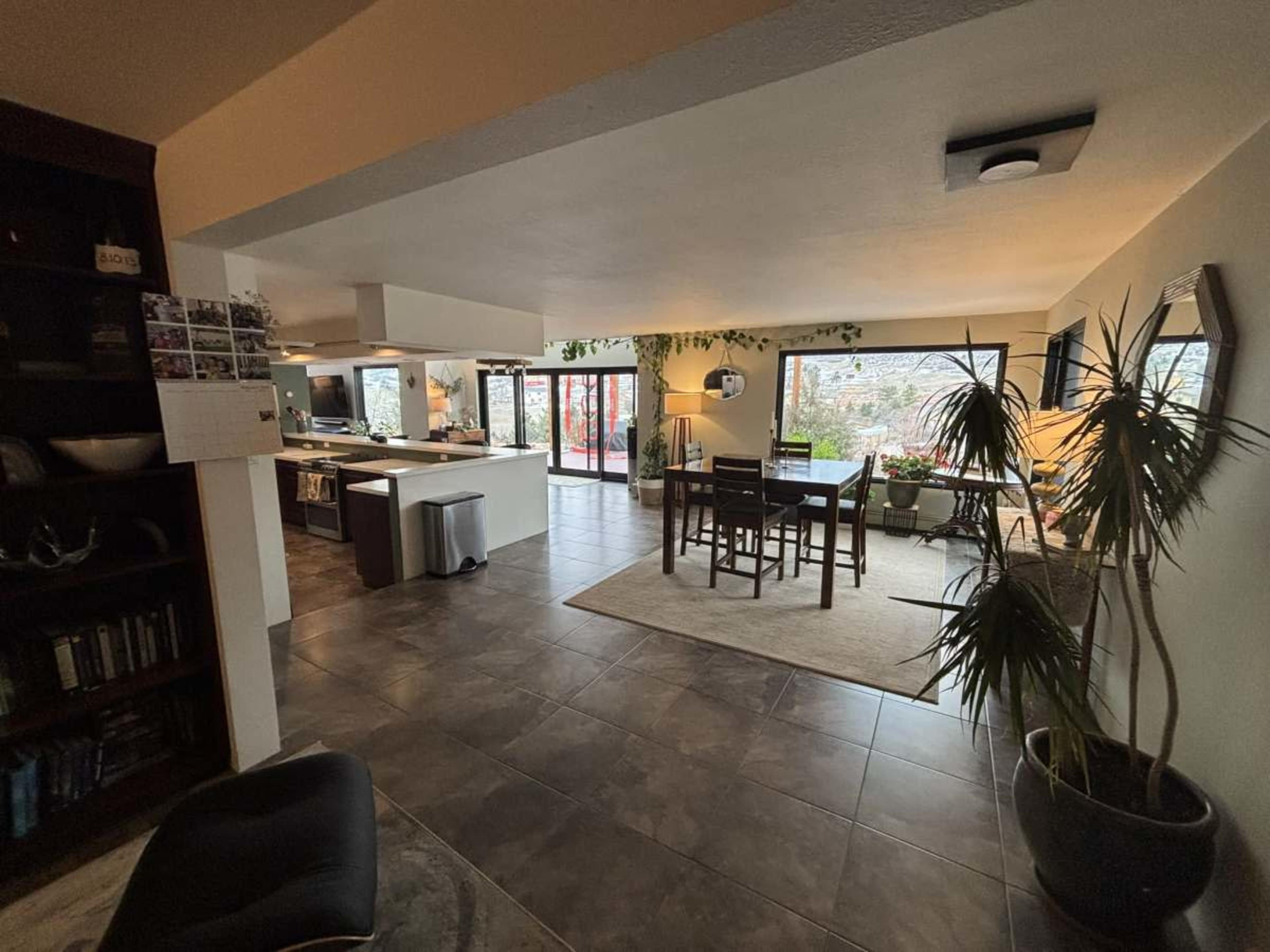 The image shows a spacious, open-concept living area with a kitchen, dining space, and large windows letting in natural light.