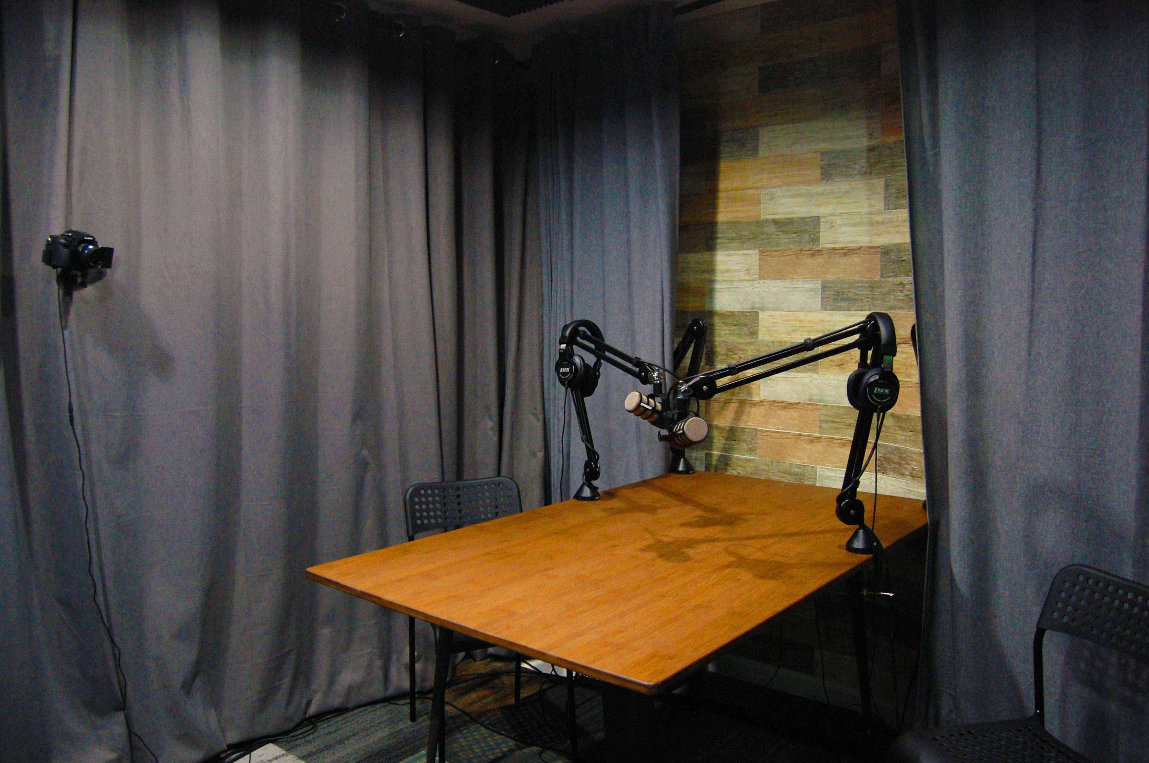 A small recording studio features a wooden table, two black chairs, and microphones mounted on adjustable arms, surrounded by dark curtains and a patterned wall.