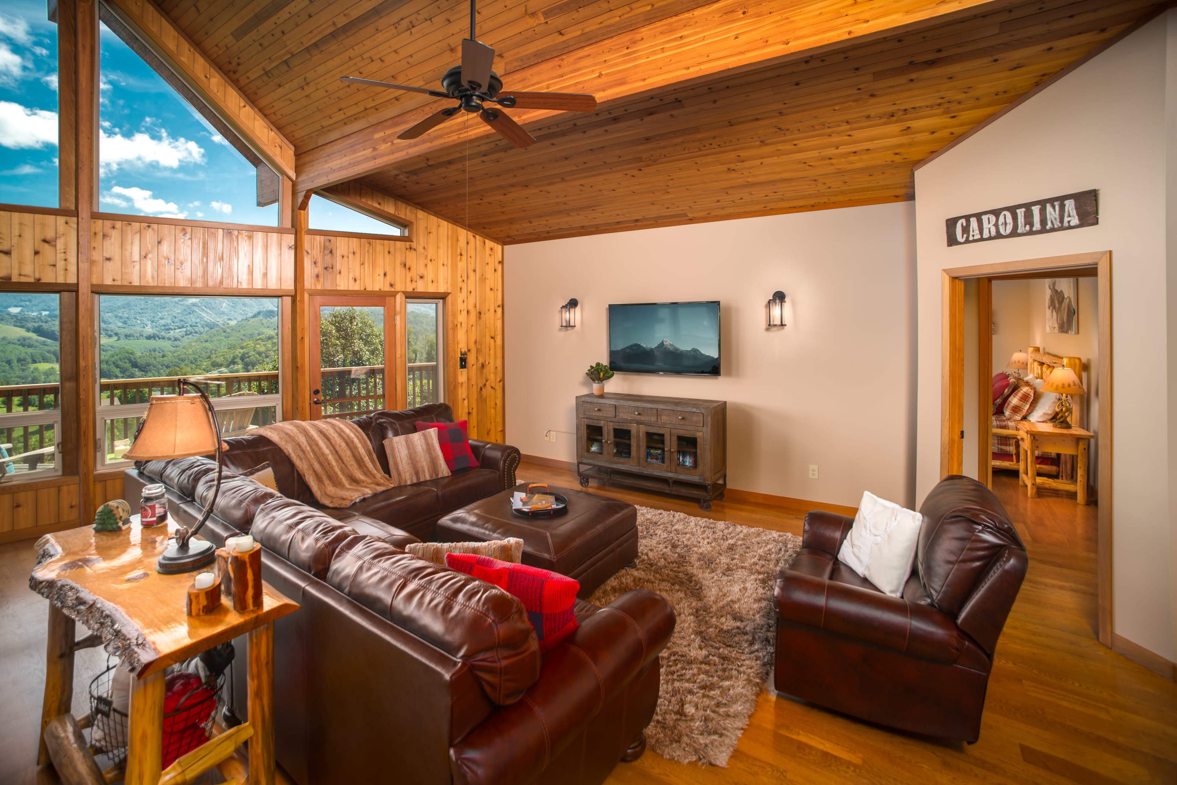 The living room features a cozy seating area with leather sofas, a wooden ceiling, and large windows offering a view of the surrounding landscape.