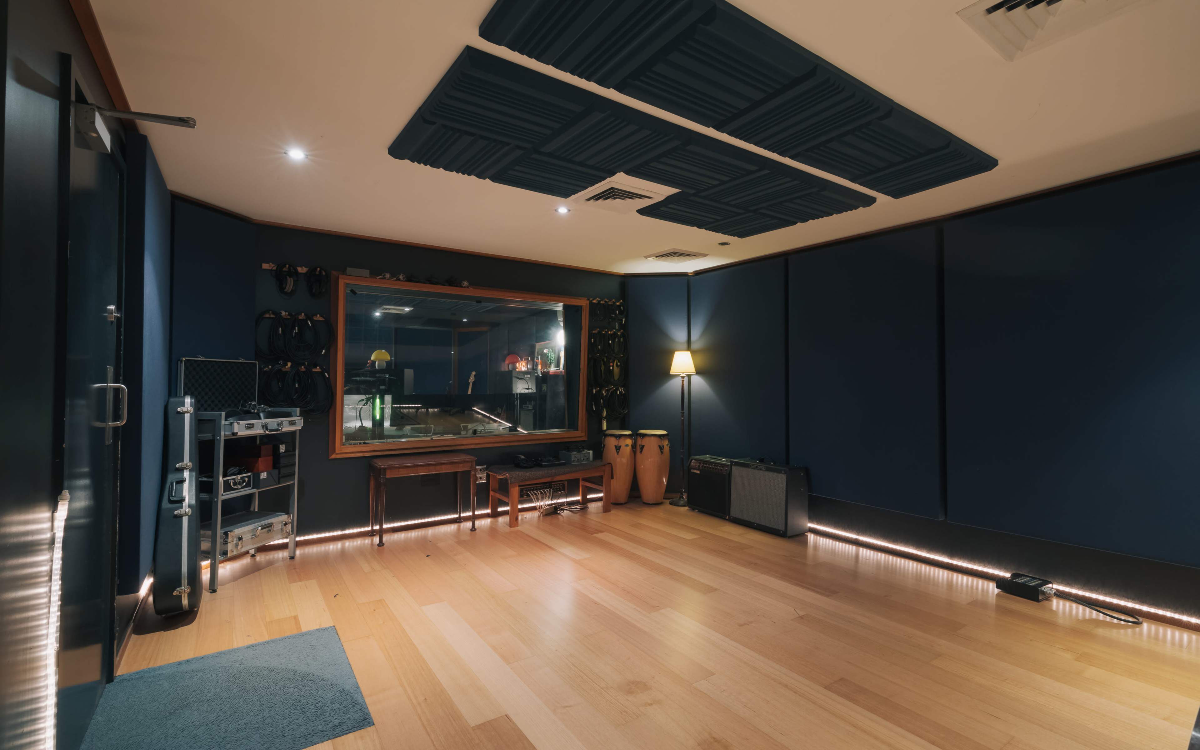 A sound studio featuring wooden floors, dark blue acoustic panels on the walls, a control window, and various musical instruments along with seating.