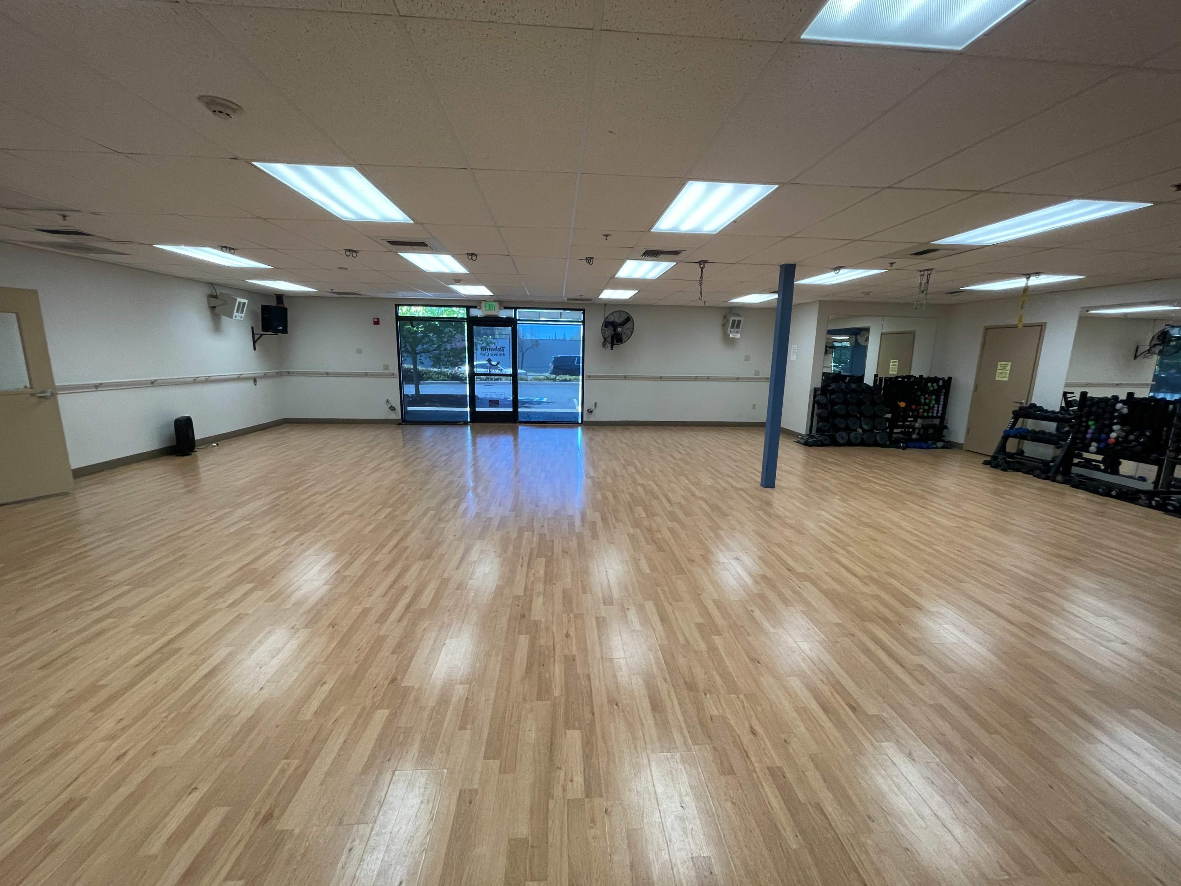 The image shows a spacious, well-lit fitness studio with wooden flooring and large windows at one end.