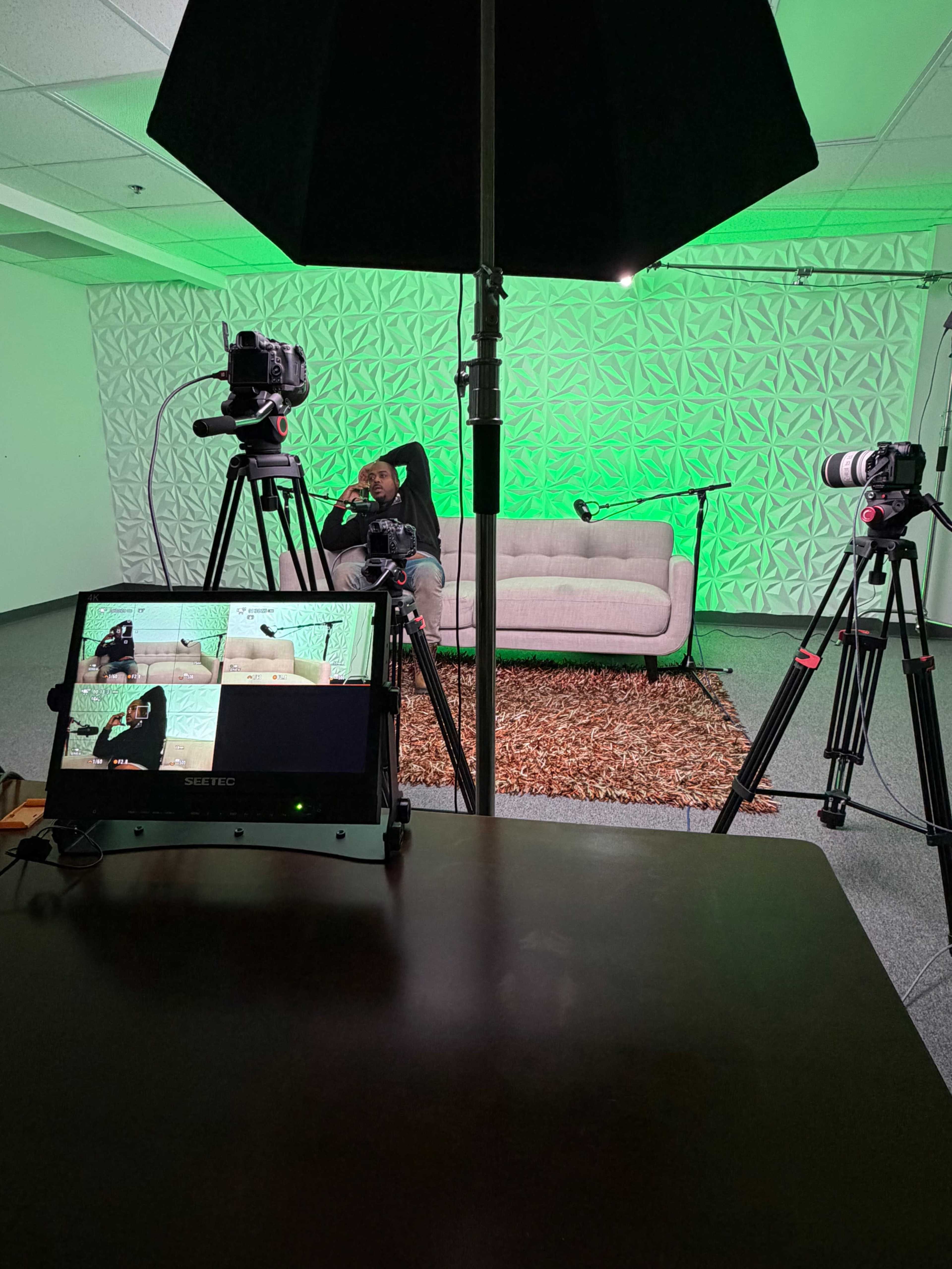 The image shows a video recording setup featuring two cameras on tripods facing a beige couch in front of a textured green backdrop, with a monitor displaying multiple camera angles on a dark table.