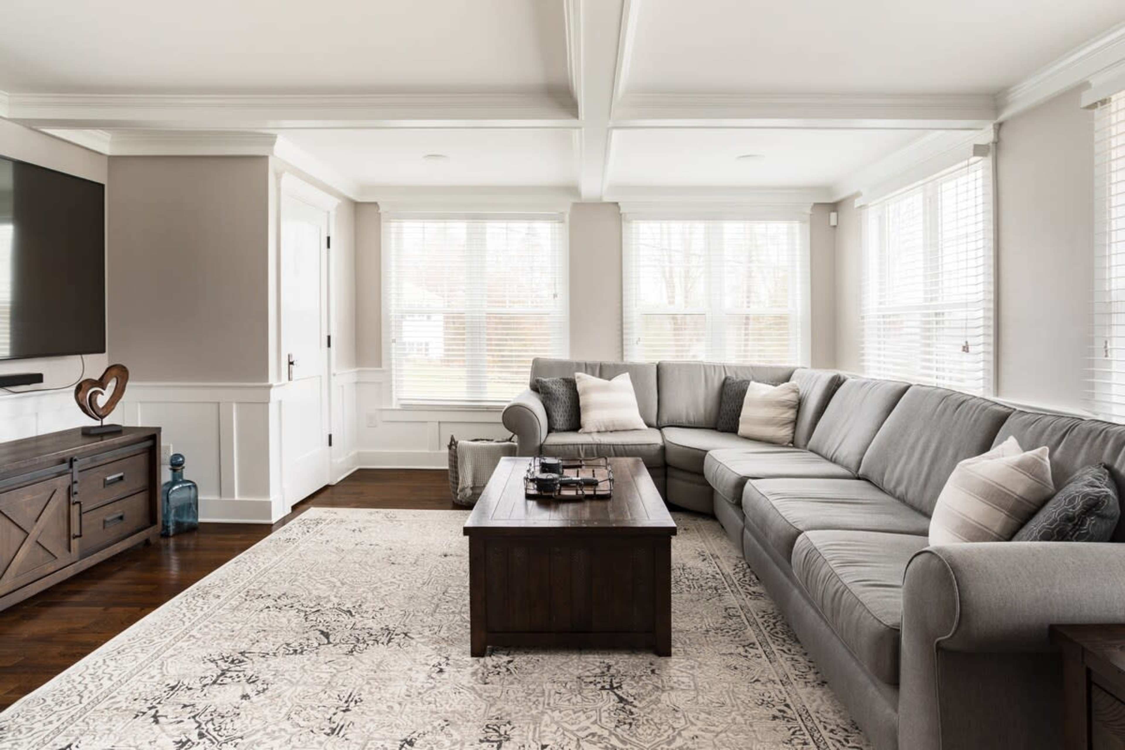 A light-filled living room features a gray sectional sofa, wooden coffee table, and a television mounted on the wall, with windows providing outdoor views.