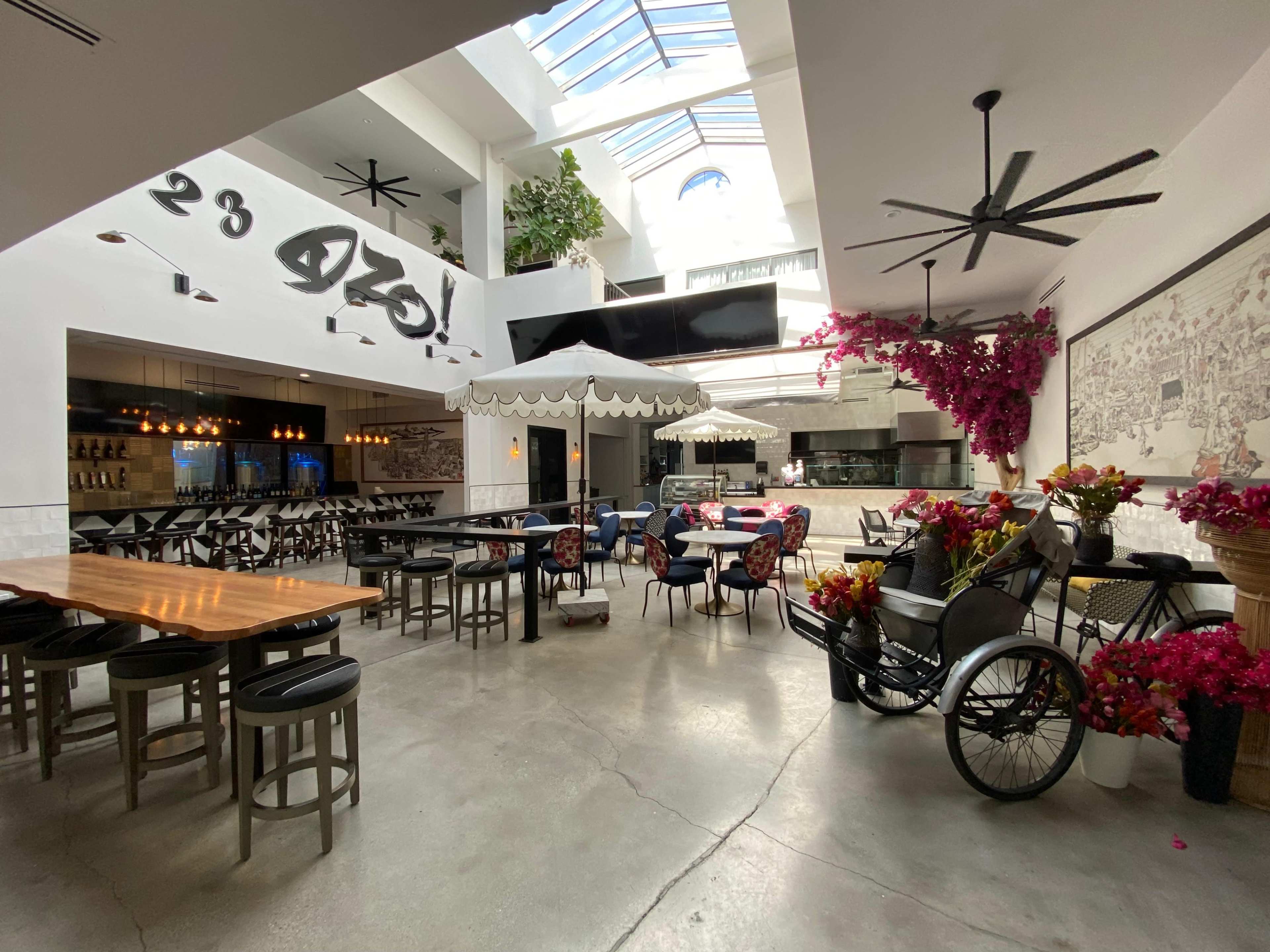 The image shows a spacious restaurant interior with modern decor, featuring a mix of seating arrangements, large windows, and decorative plants.