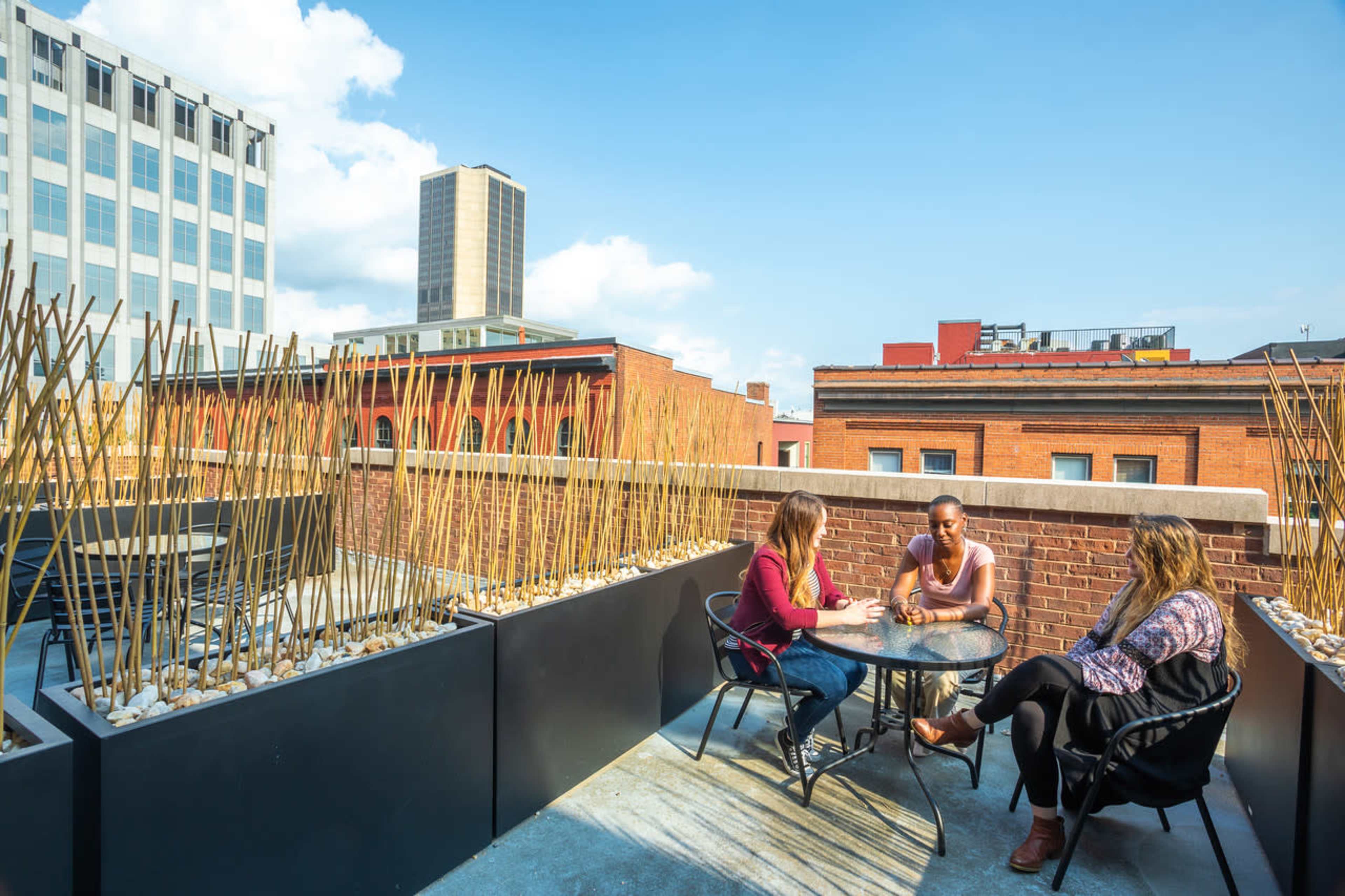 Three people are seated at a round table on a rooftop patio surrounded by modern architecture and tall plants.