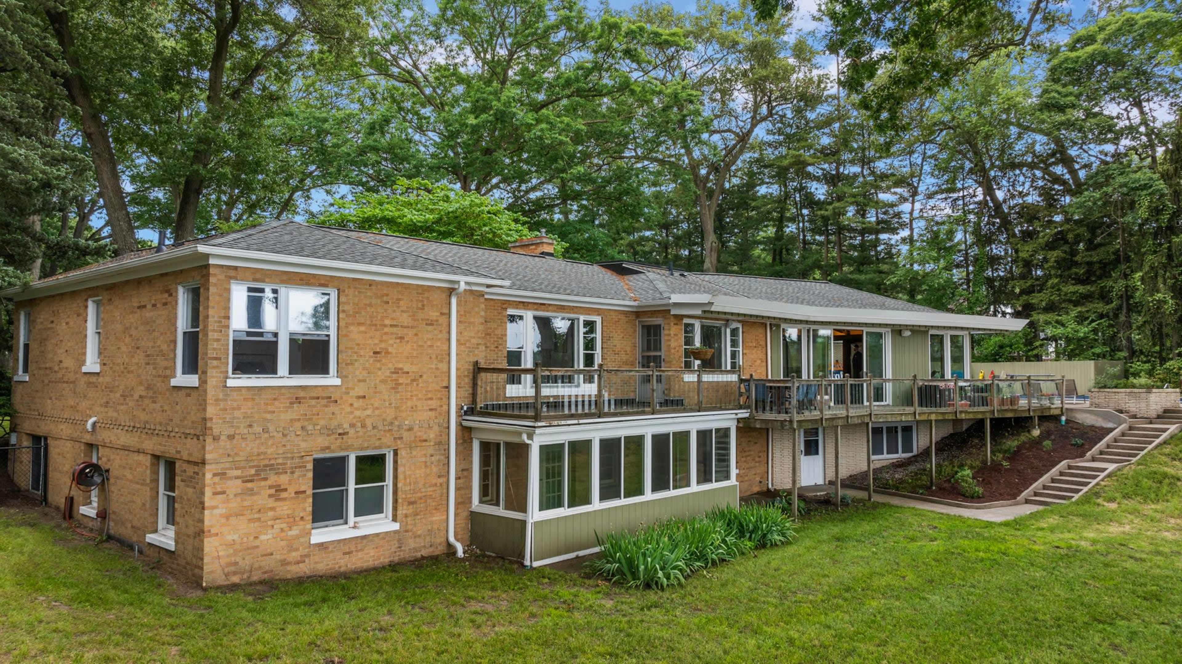 The image shows a two-story brick house with large windows, a deck, and a landscaped backyard surrounded by trees.