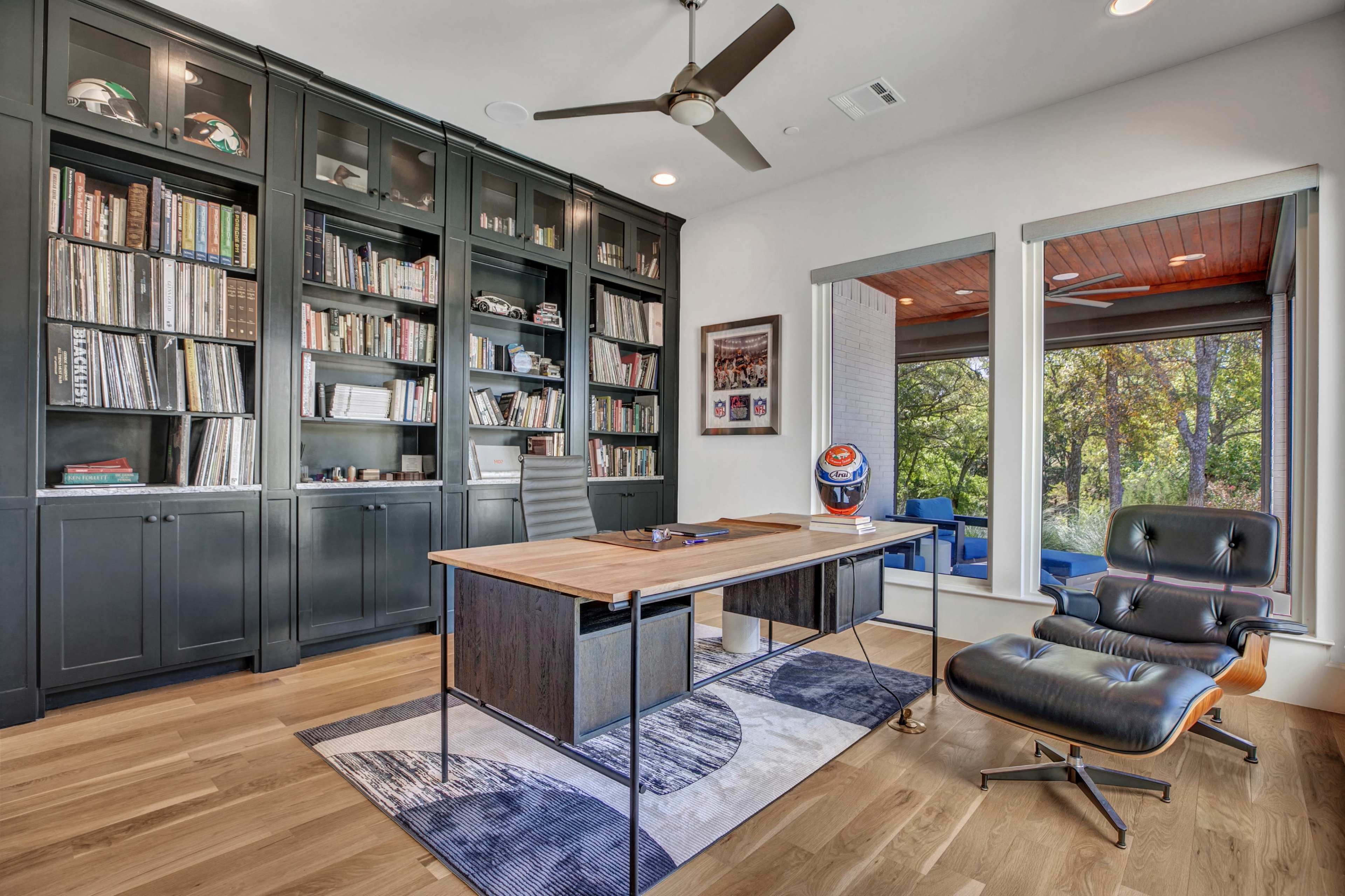 A modern office space featuring a large wooden desk, a black leather chair, and a wall-mounted bookshelf filled with books, alongside a window with a view of greenery outside.