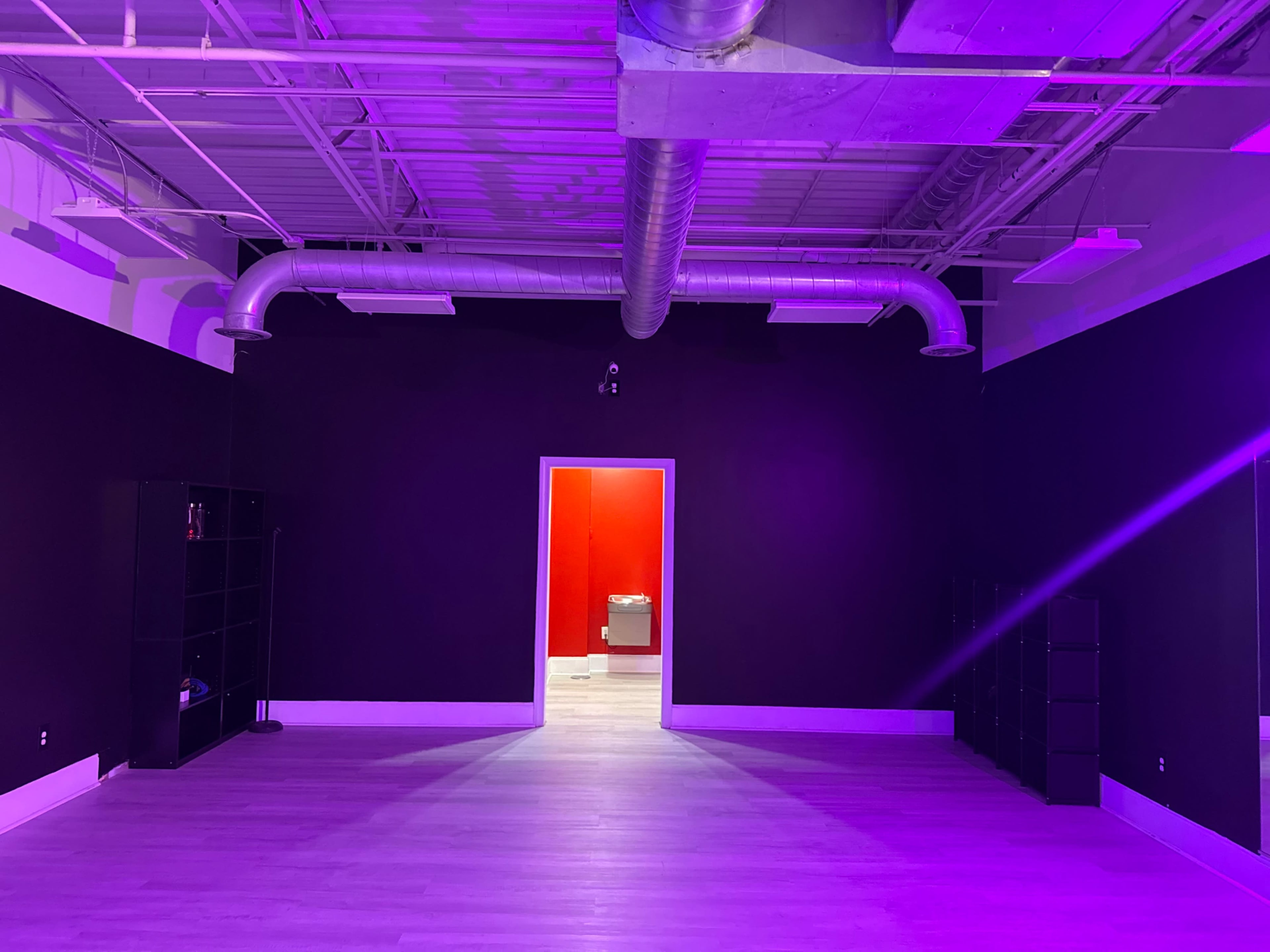 The image shows an empty room with dark walls and purple lighting, featuring a red door on one side and shelves against the walls.
