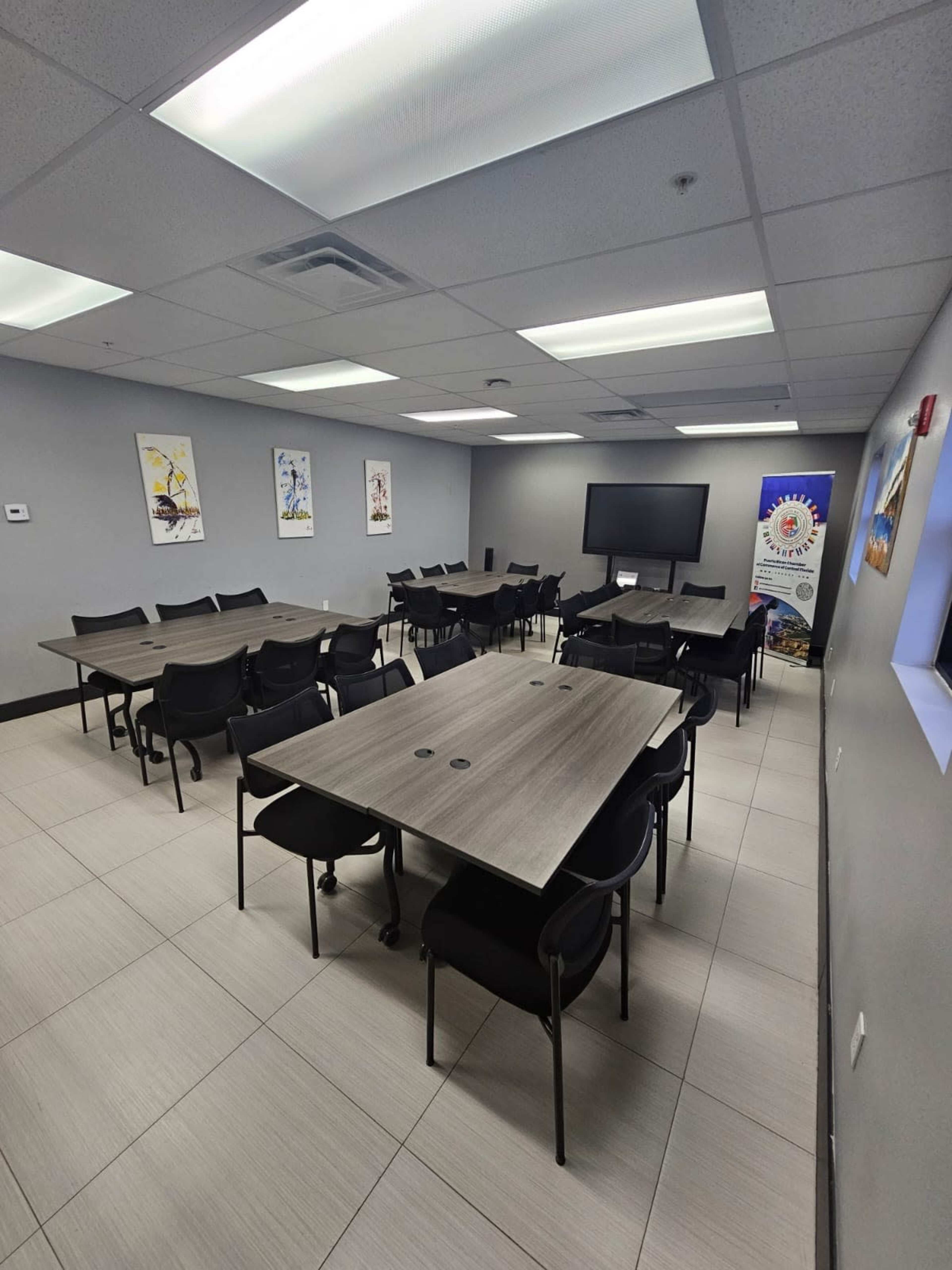 The image shows a conference room with multiple rectangular tables and black chairs arranged around them, alongside a large screen and decorative wall posters.