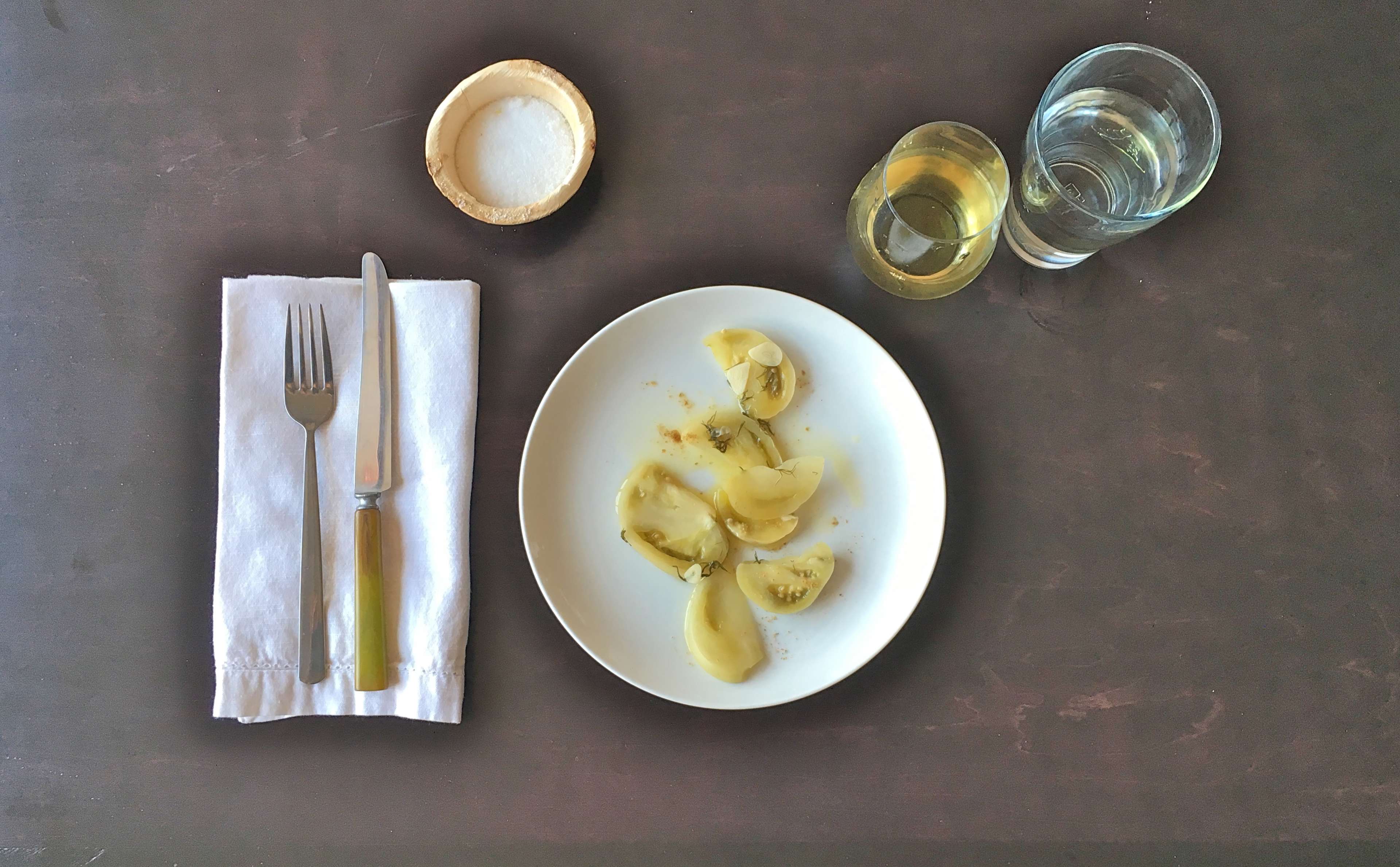 The image shows a table setting with a white plate of pasta, a bowl of salt, a wine glass, and a glass of water, accompanied by a fork and knife on a white napkin.