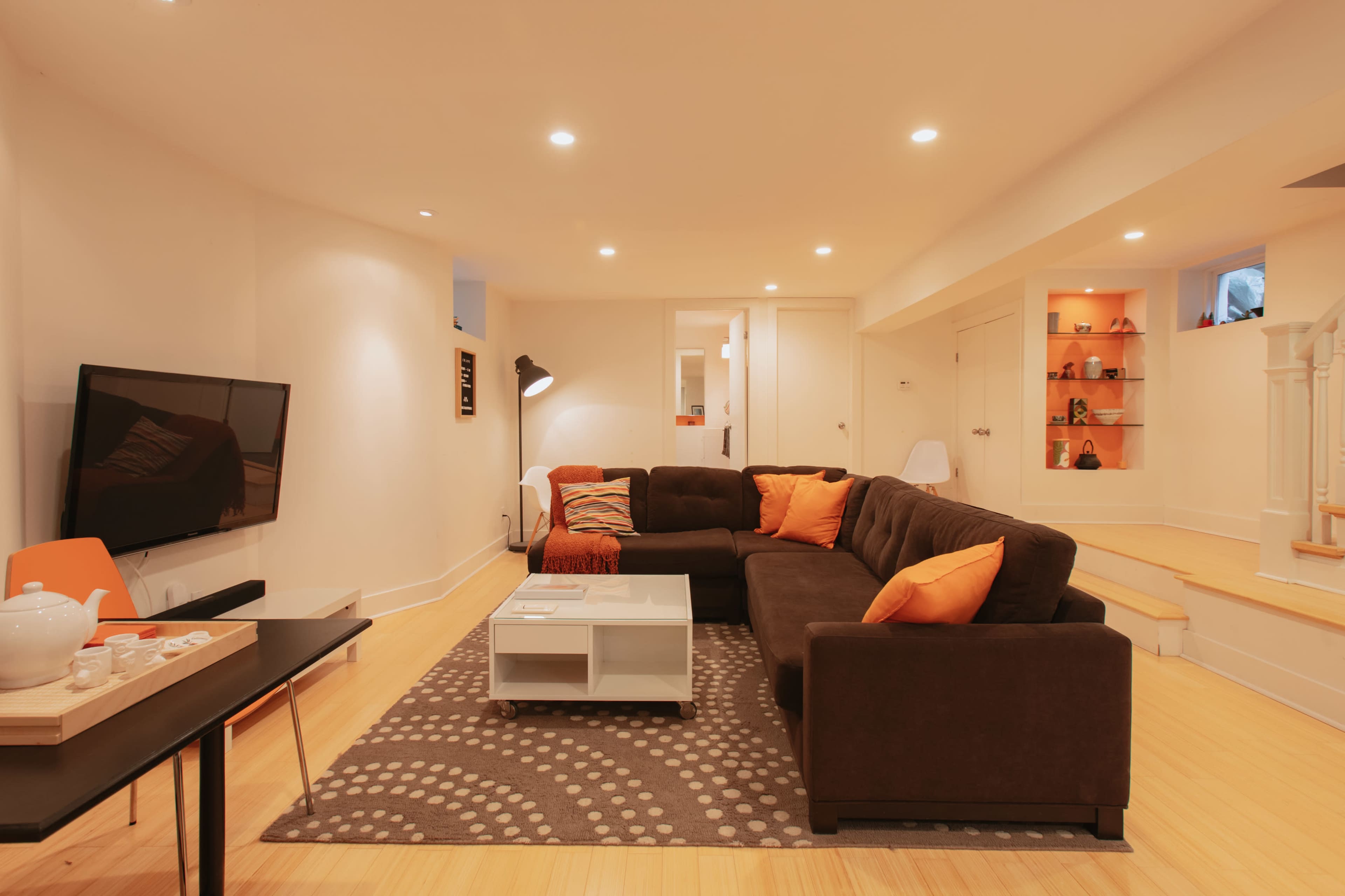 A well-lit basement living room featuring a dark sectional sofa with orange pillows, a white coffee table, a flat-screen TV, and a light-colored staircase.