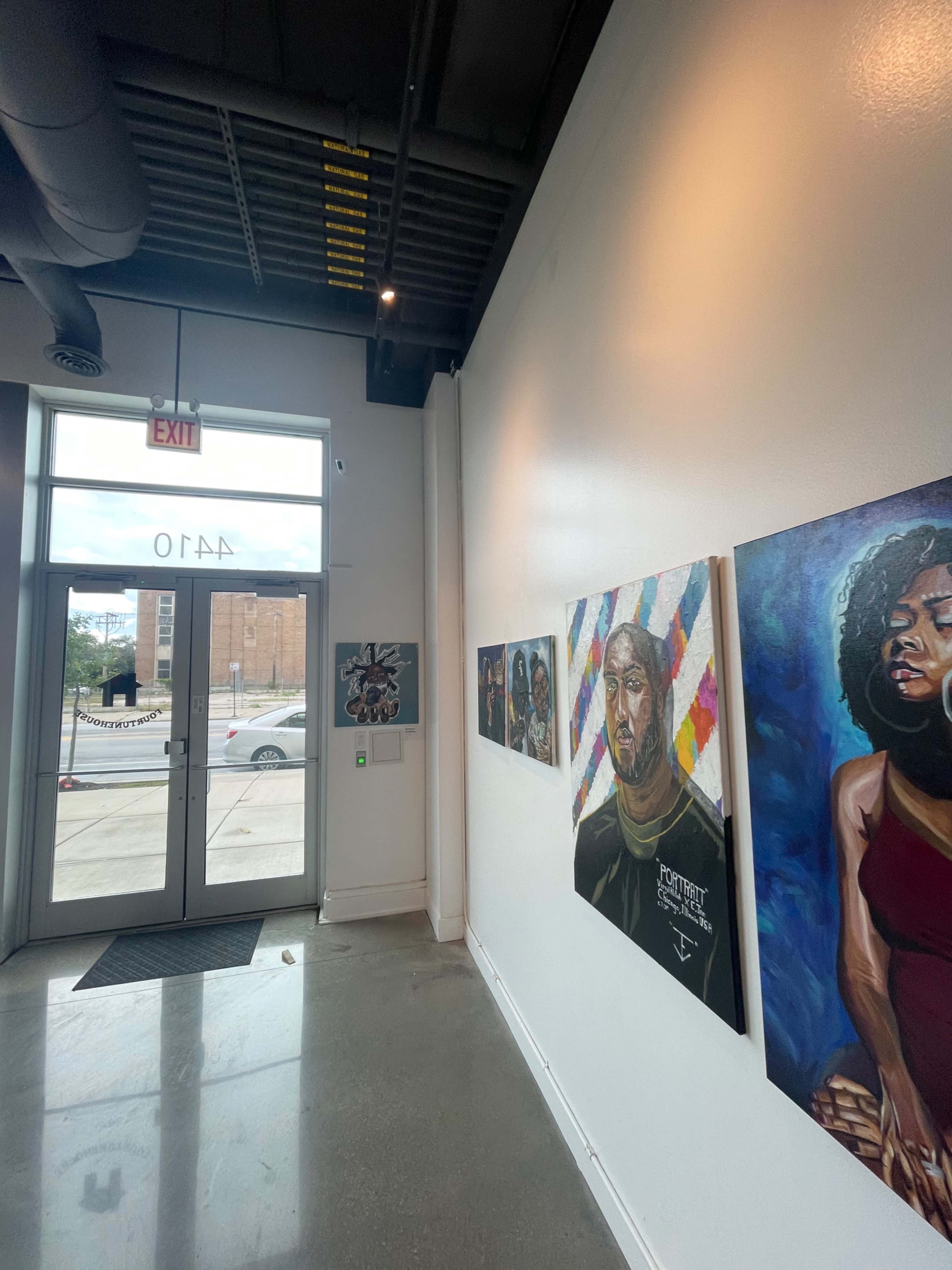 A gallery interior with a gallery wall displaying several colorful paintings, and a doorway leading outside.
