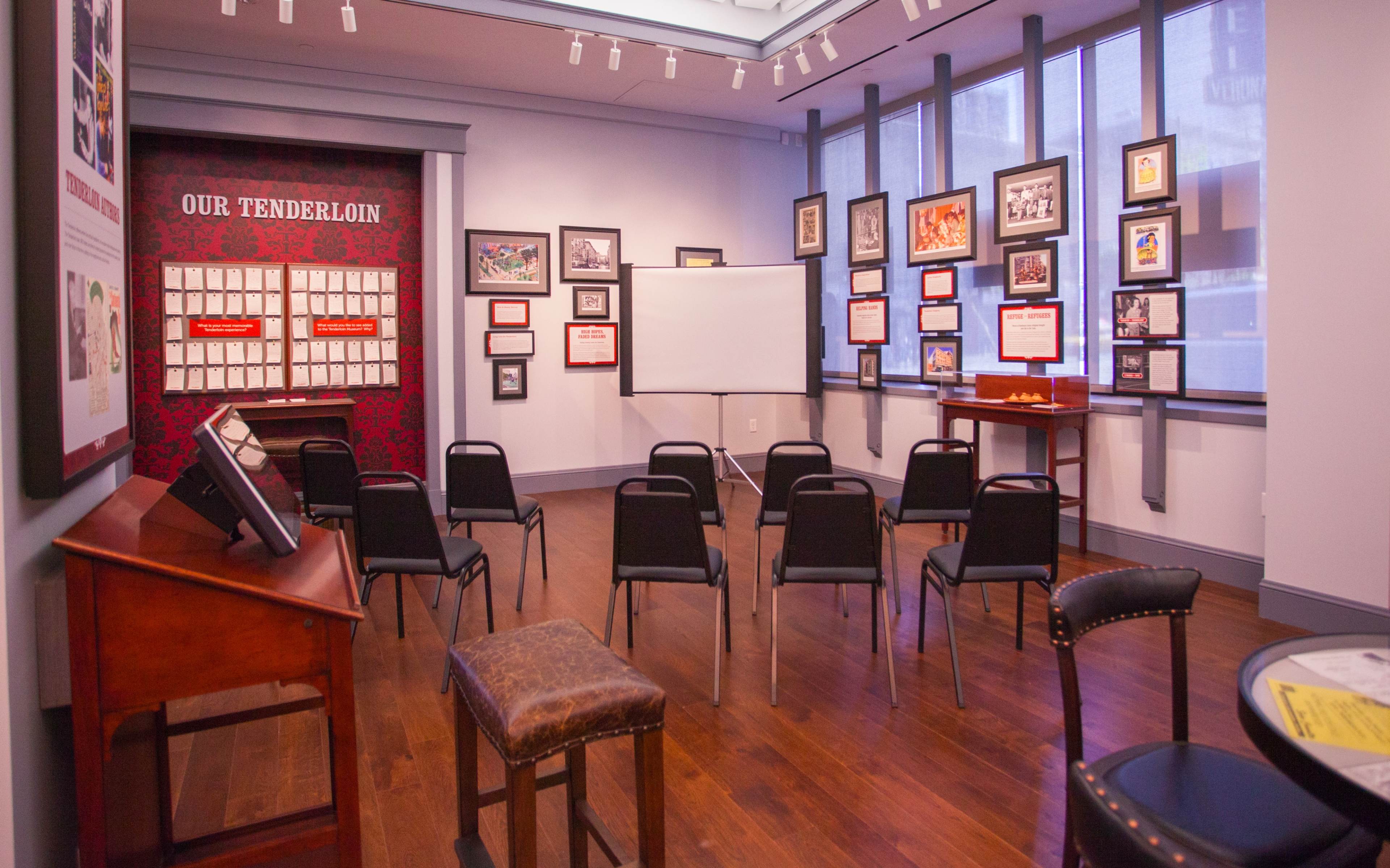 A meeting room with chairs arranged in rows facing a blank projection screen, surrounded by framed photographs and information displays on the walls.