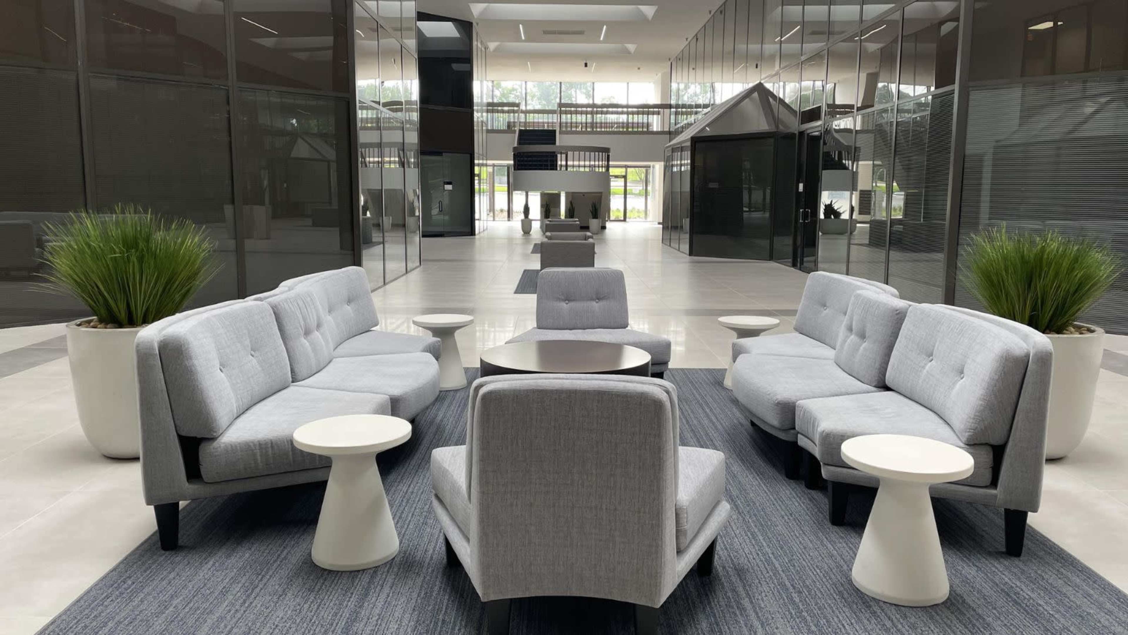 A modern lounge area with gray sofas, white side tables, and decorative plants, set in a spacious, glass-walled interior.