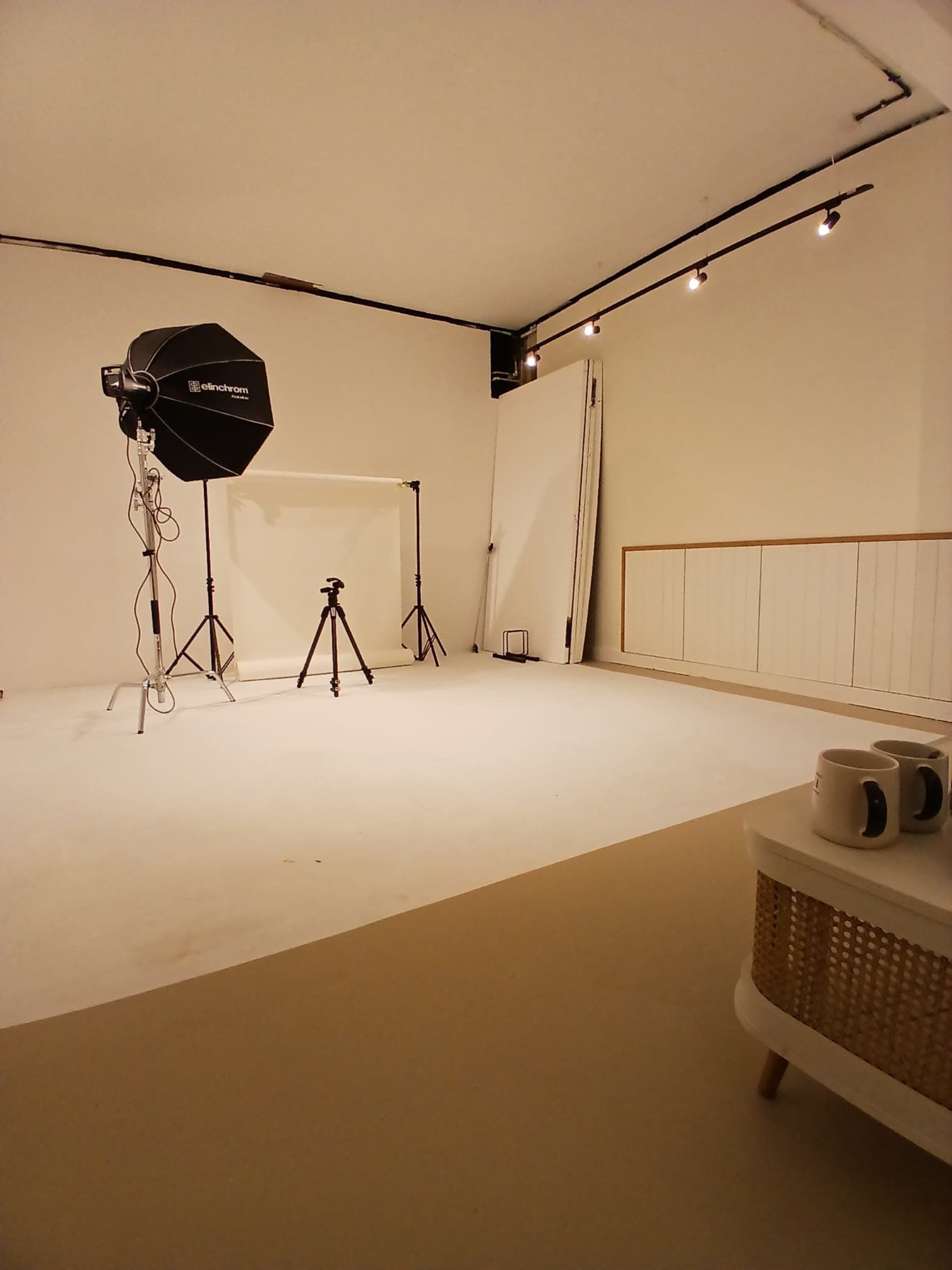 The image depicts a photography studio featuring a light source, a backdrop setup, a camera tripod, and a table with mugs in a minimalist environment.