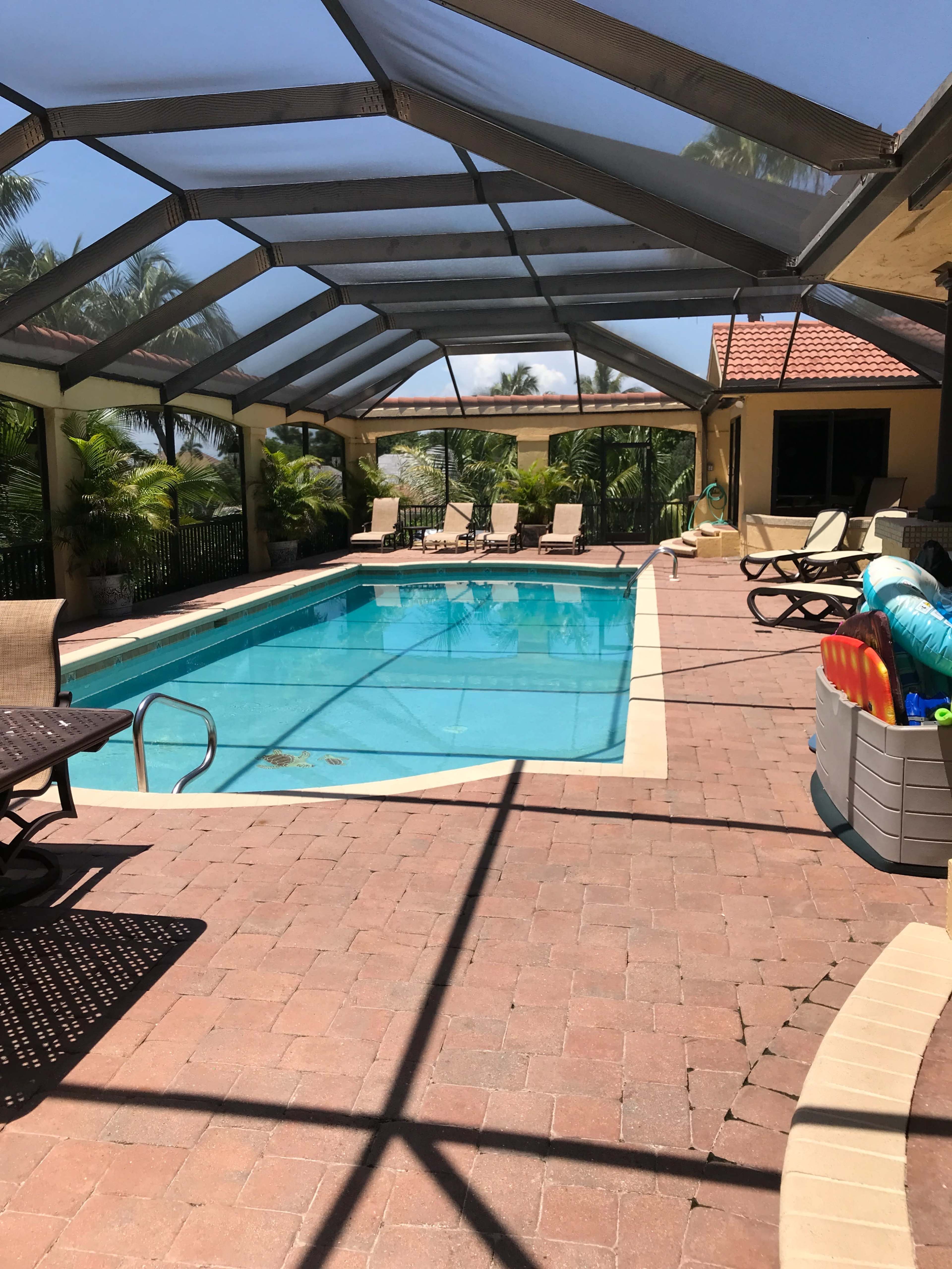 A rectangular swimming pool is enclosed by a screened patio, surrounded by lounge chairs and tropical plants.