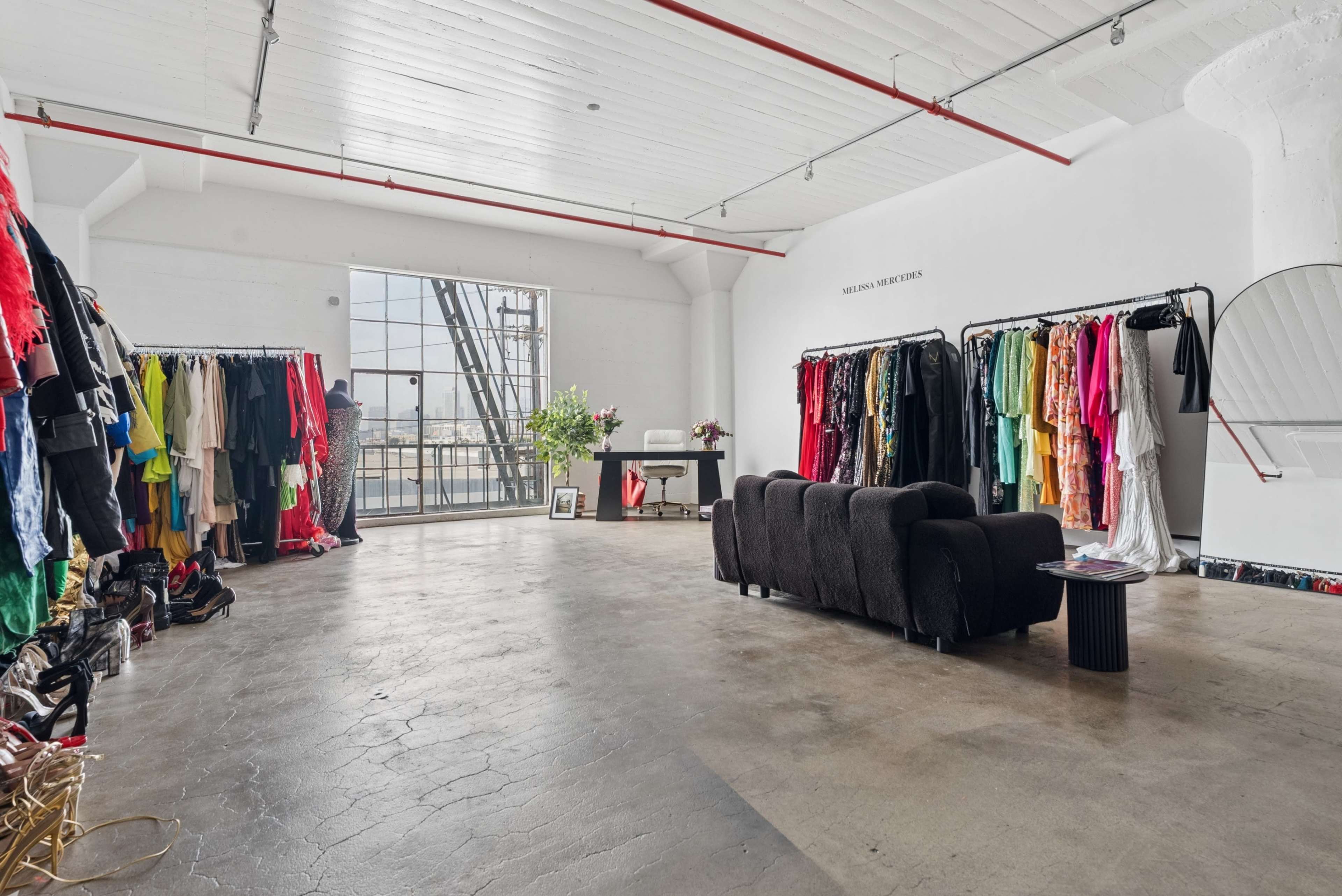 The image depicts a spacious interior with clothing racks filled with various garments and a black couch positioned nearby under natural light from large windows.