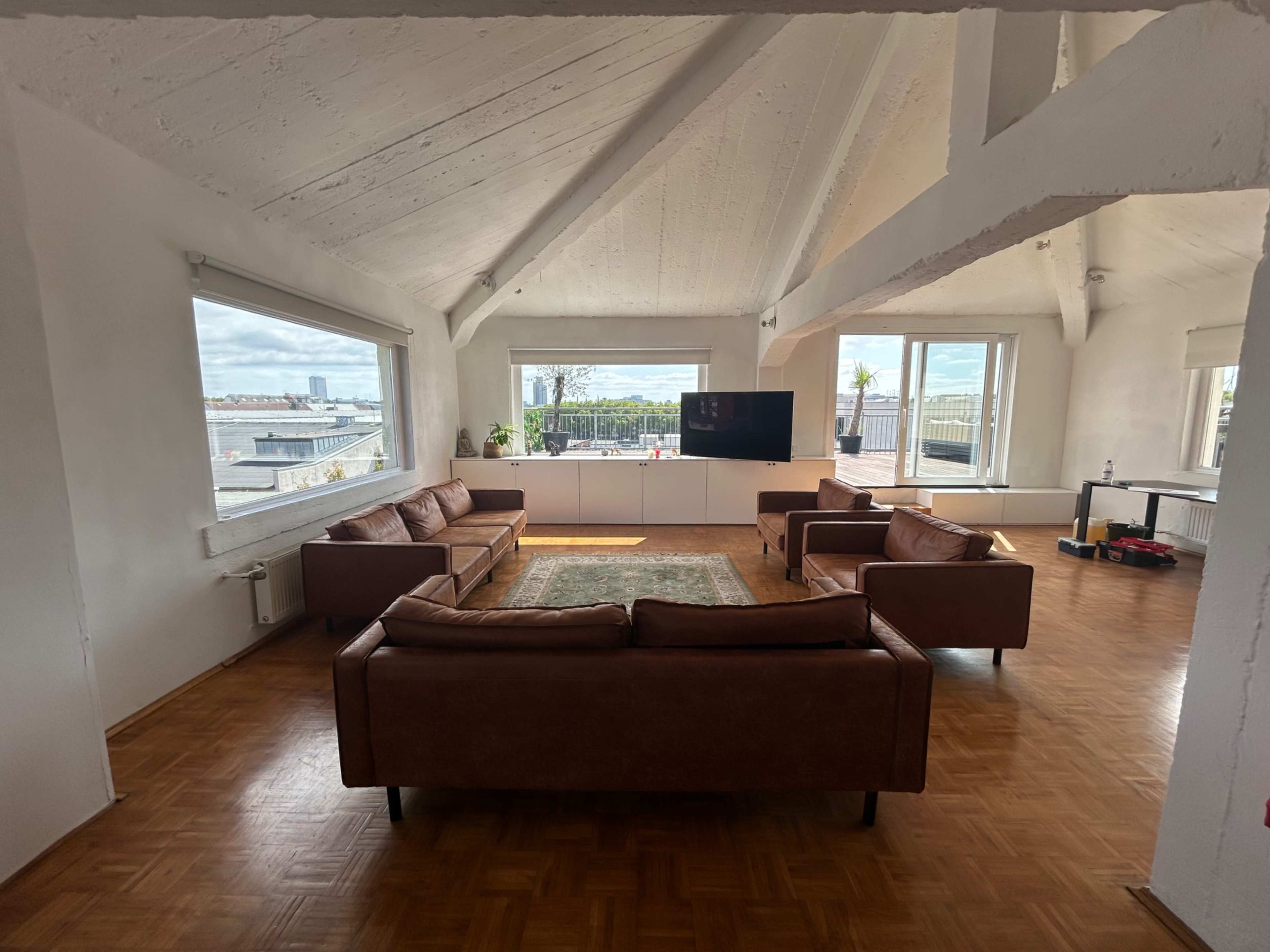 The image shows a spacious living room with a wooden floor, two sets of brown leather sofas arranged around a central rug, large windows providing natural light, and a television mounted on the wall.