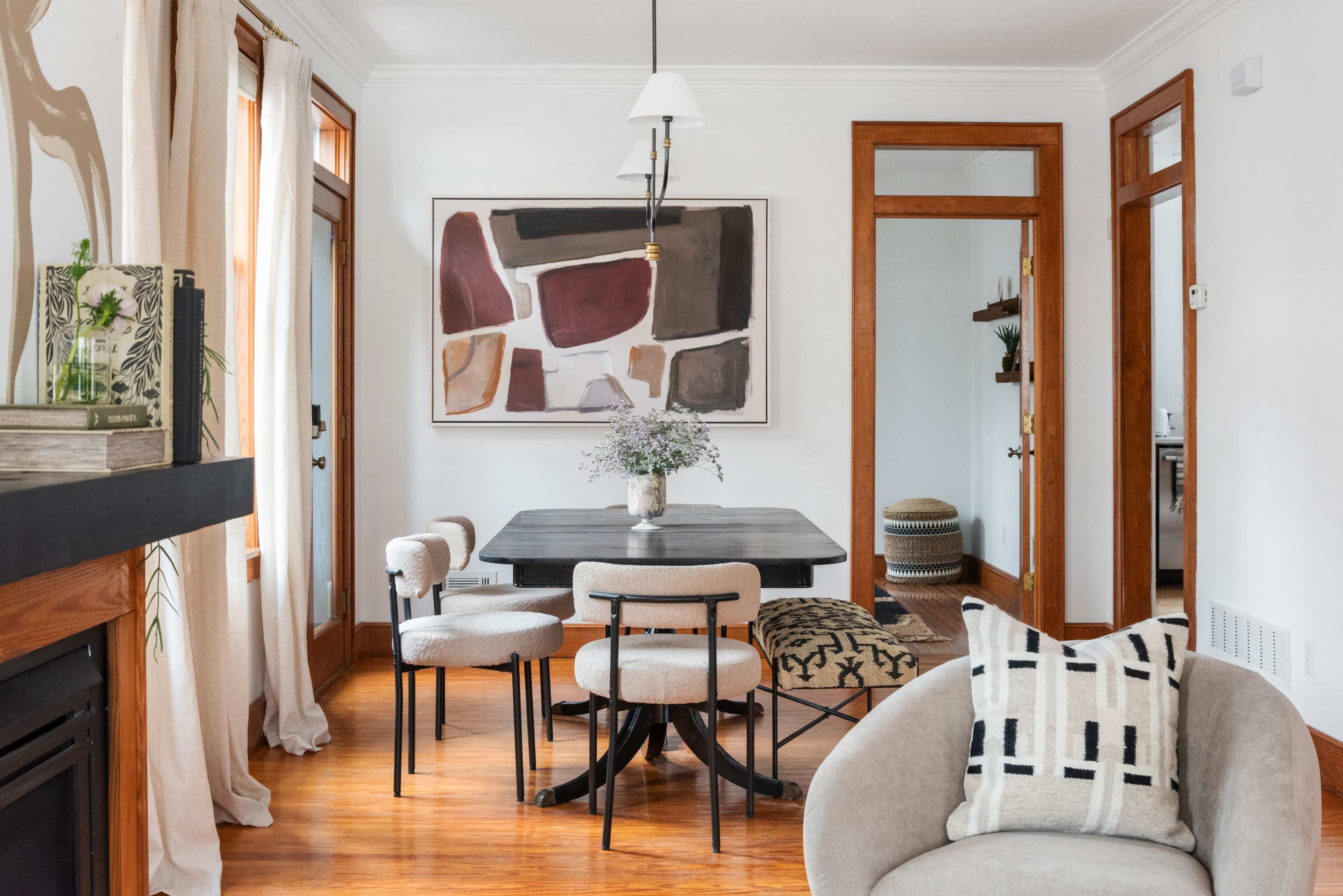 A bright dining area featuring a rectangular black dining table surrounded by upholstered chairs, with a large abstract artwork on the wall and a cozy armchair in the foreground.