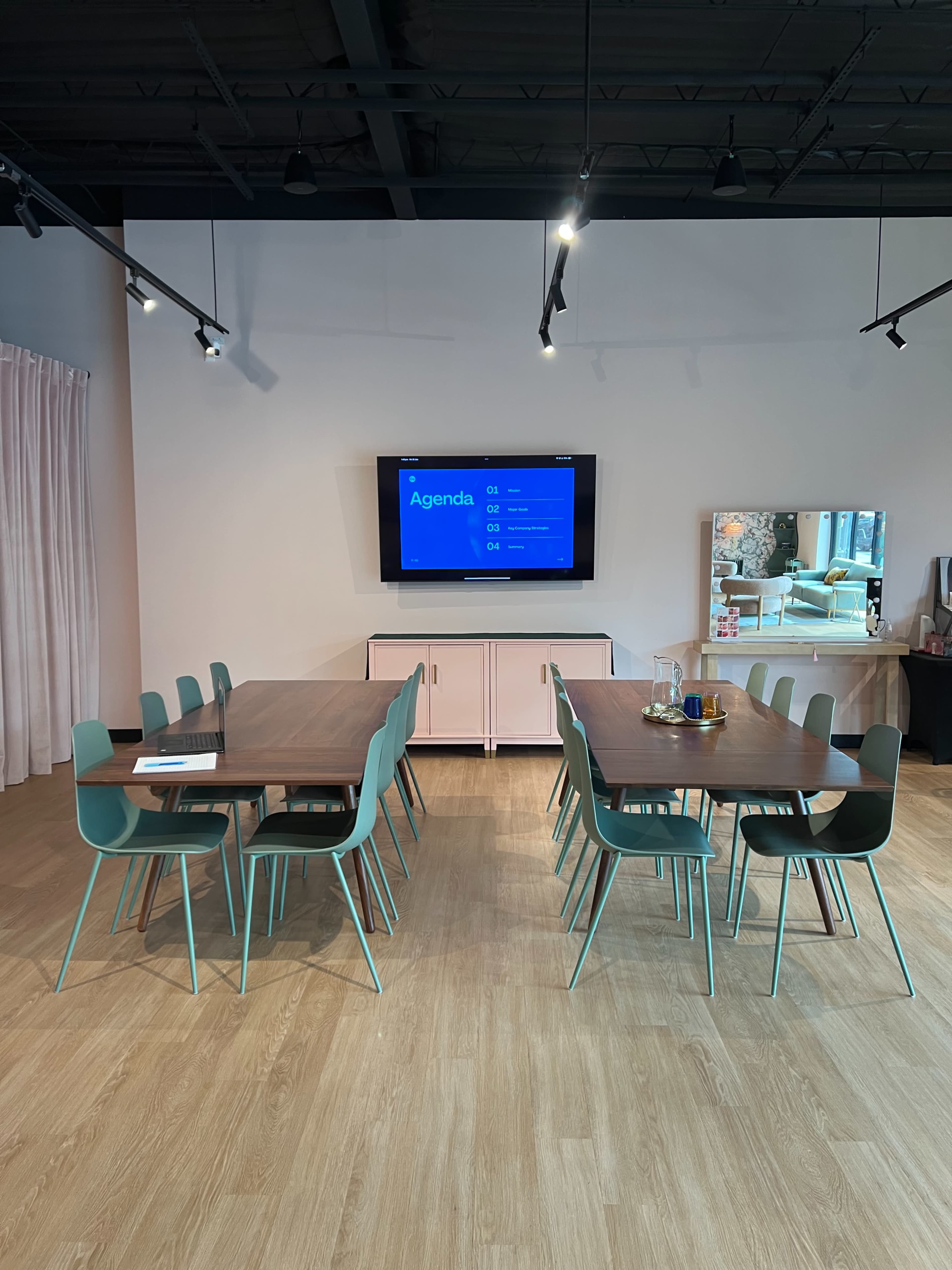 A modern conference room features two tables arranged in a U-shape, with green chairs and a wall-mounted TV displaying an agenda.