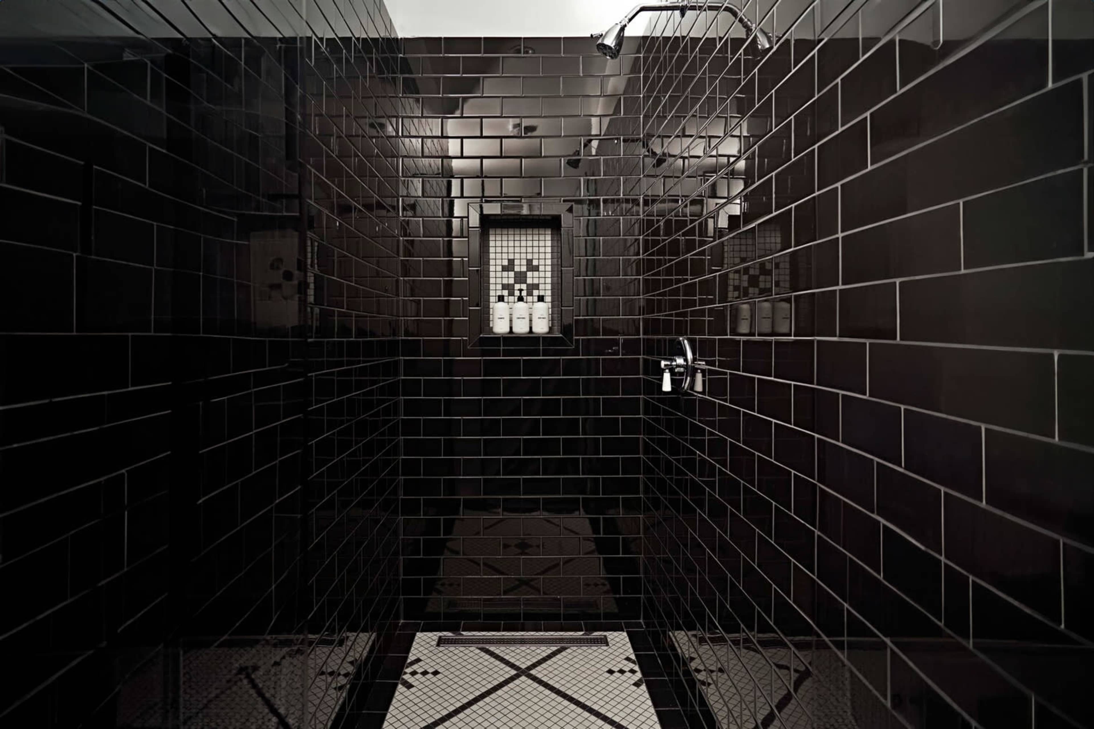 The image shows a modern shower space with black tiled walls and a patterned floor, featuring a glass shelving unit for toiletries.
